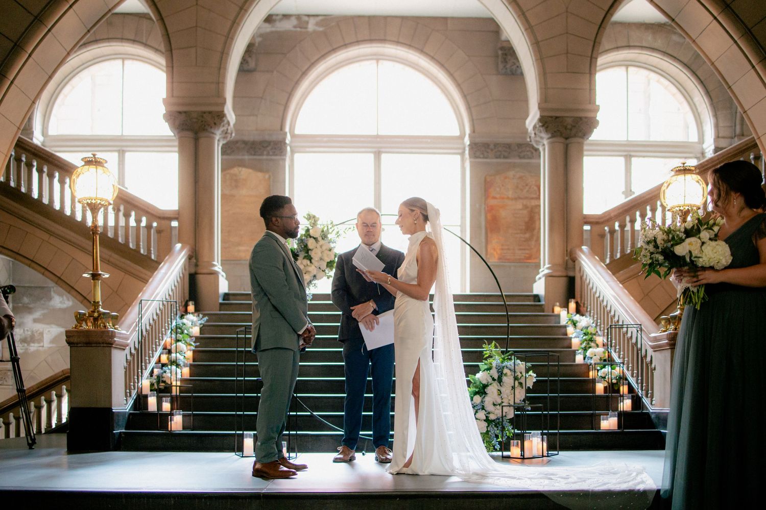 Bride and groom exchange vows at elegant indoor wedding ceremony beneath grand arched staircase.