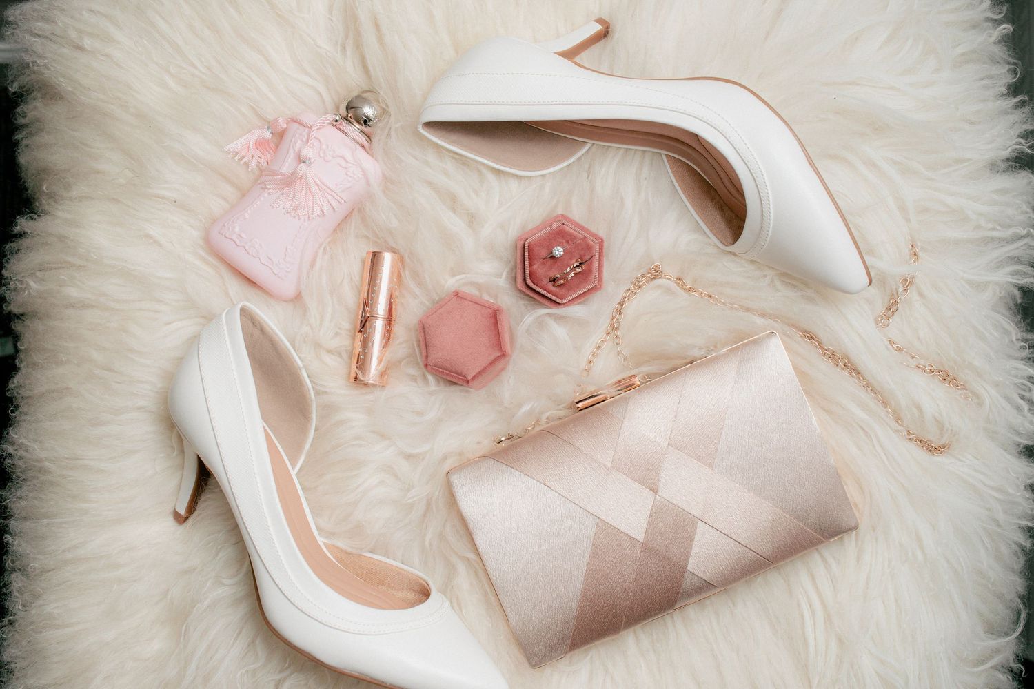 Bridal flat lay featuring white heels, pink clutch, lipstick, perfume, and hexagonal ring box on fur.