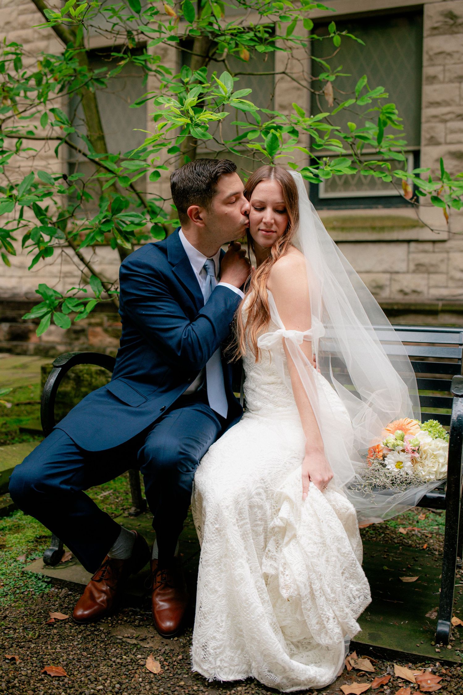 Groom in navy suit kisses bride on cheek as they sit on bench in lush garden setting.