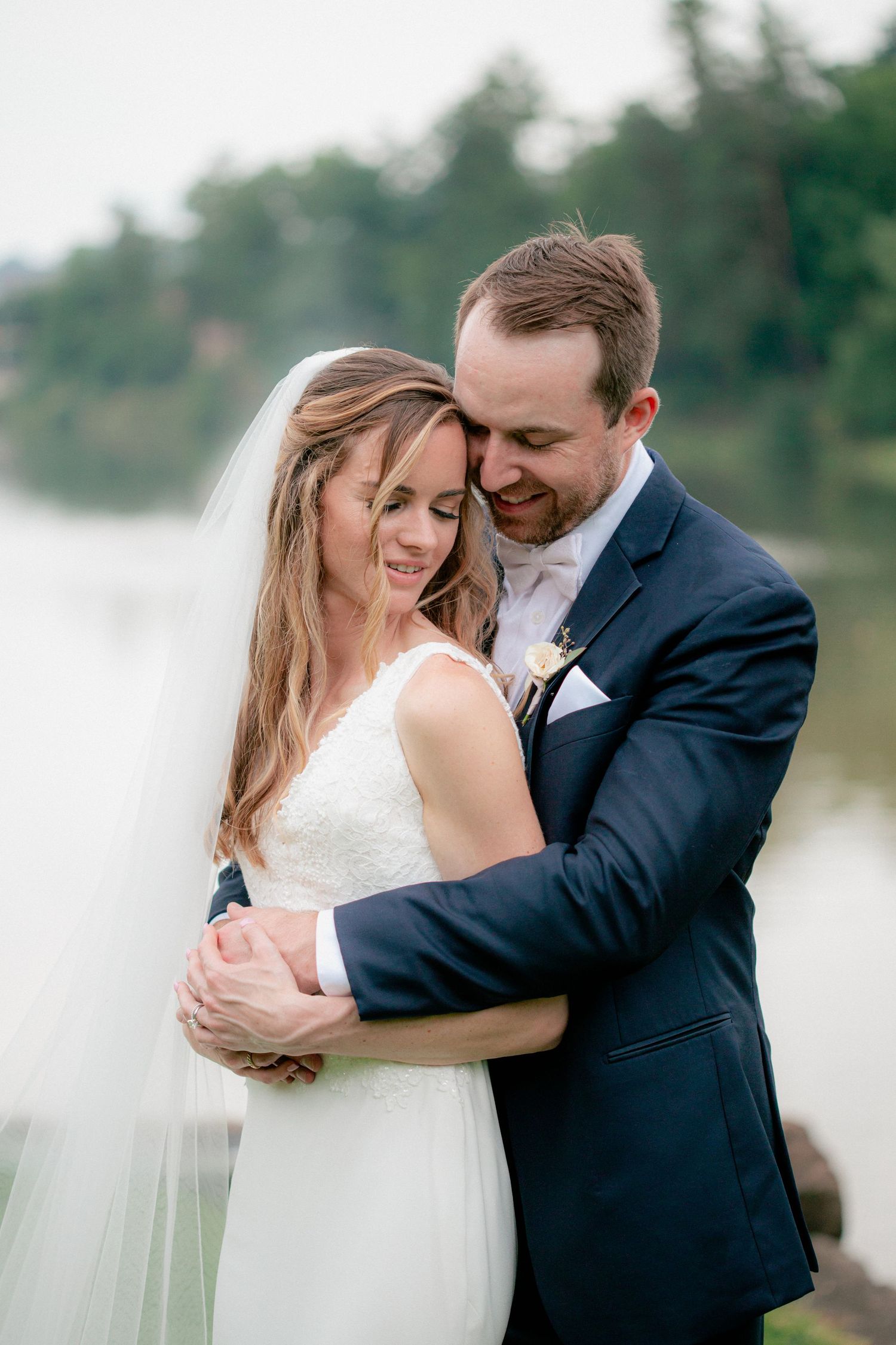 Groom embracing bride from behind near misty lakeside, both smiling in a tender wedding portrait.