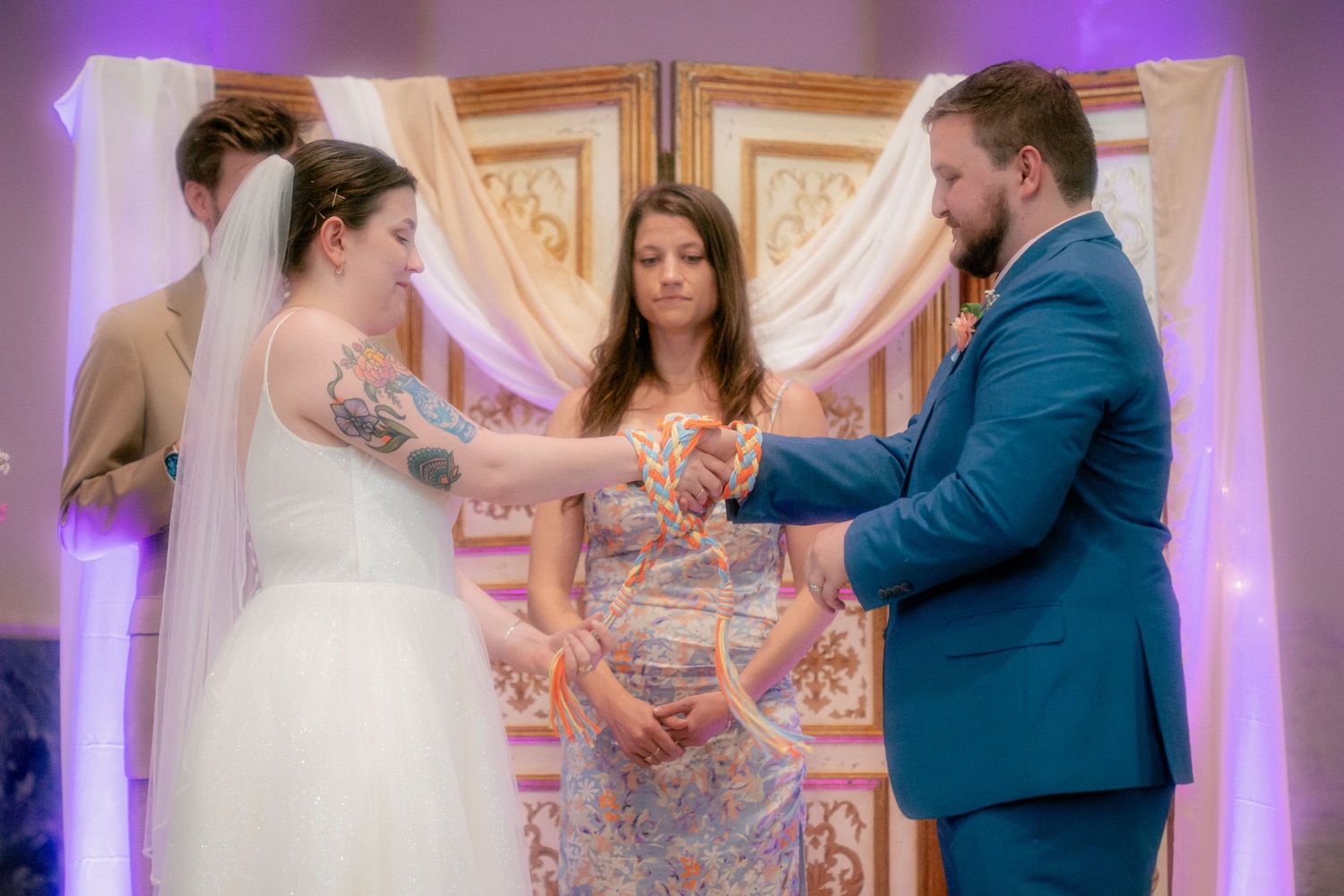 Bride and groom exchange vows at wedding ceremony with officiant, bride wearing white gown and veil, groom in blue suit.