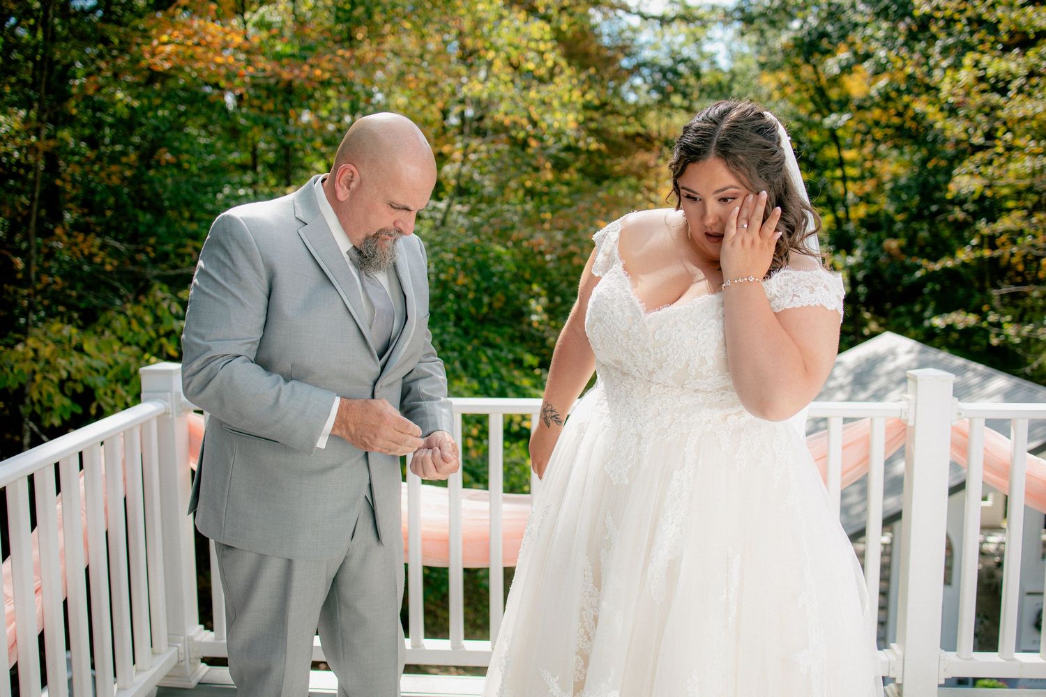 Emotional bride wipes tears during outdoor first look with father in gray suit on white bridge.