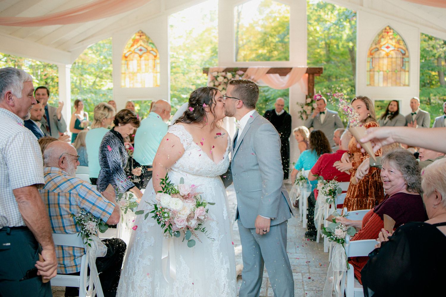 Newlyweds sharing a kiss while guests celebrate with flower petals under an outdoor wedding pavilion.