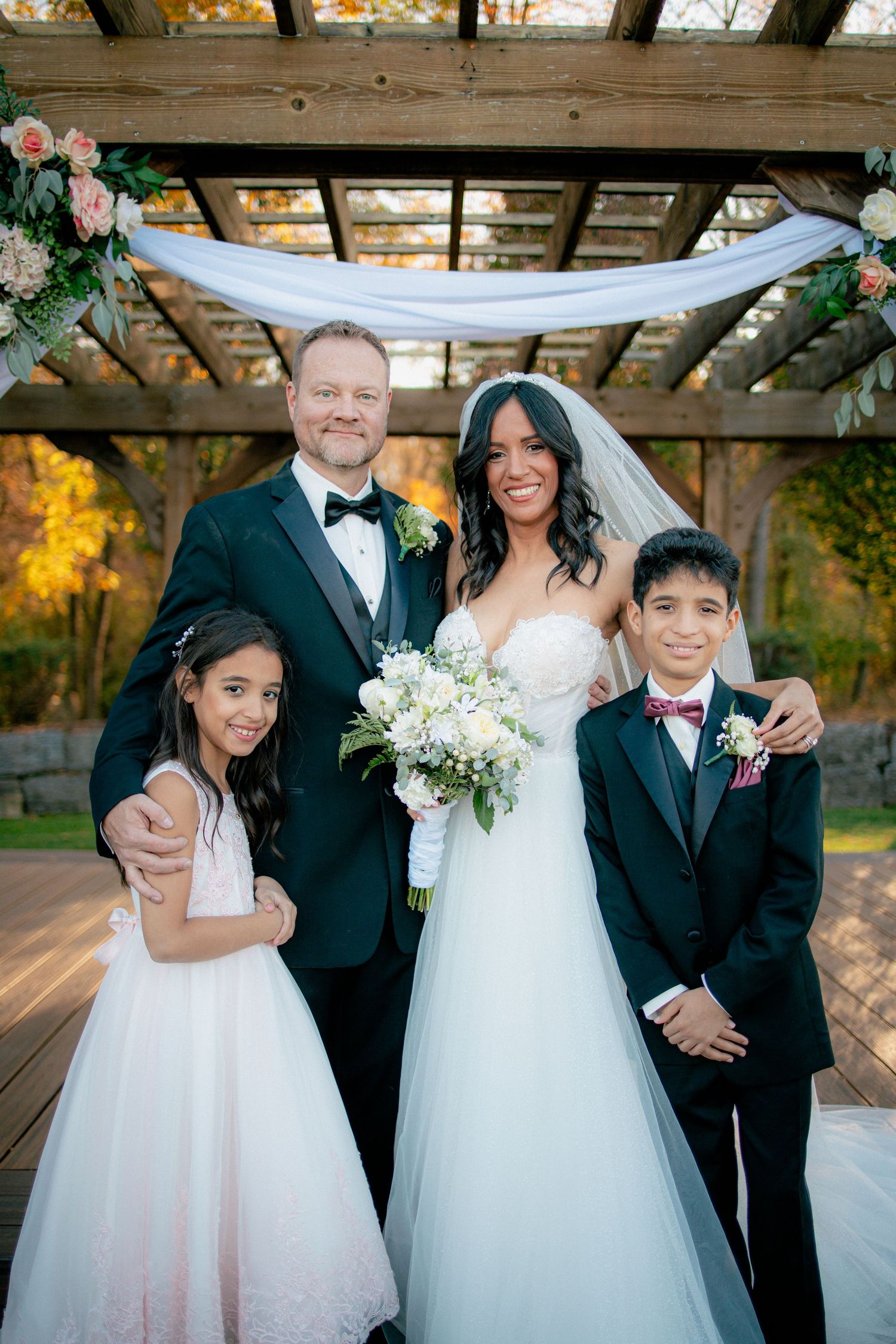 Bride and groom posing with two children under a wooden pergola decorated with white floral draping.