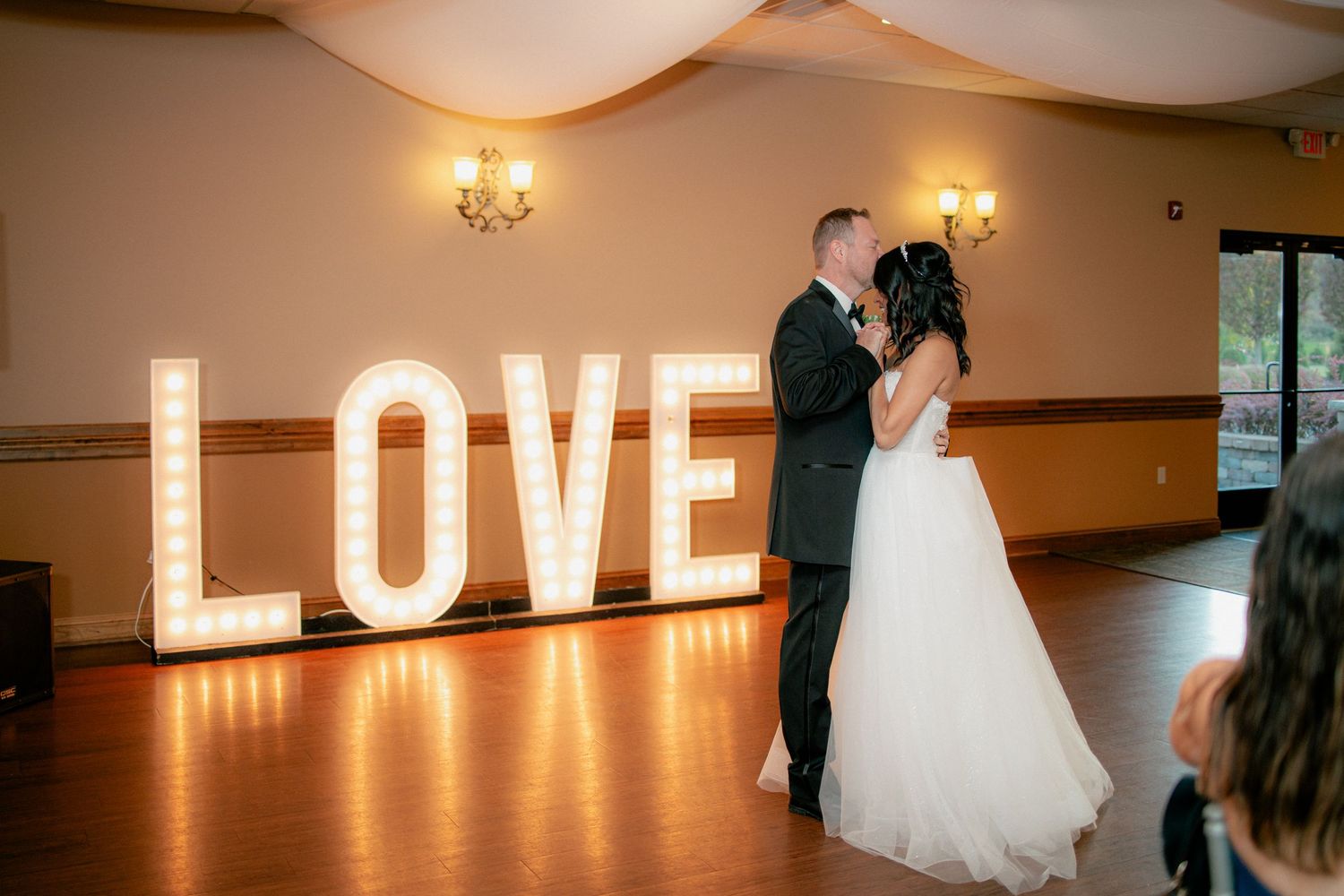 Bride and groom share first dance in front of illuminated LOVE marquee letters.