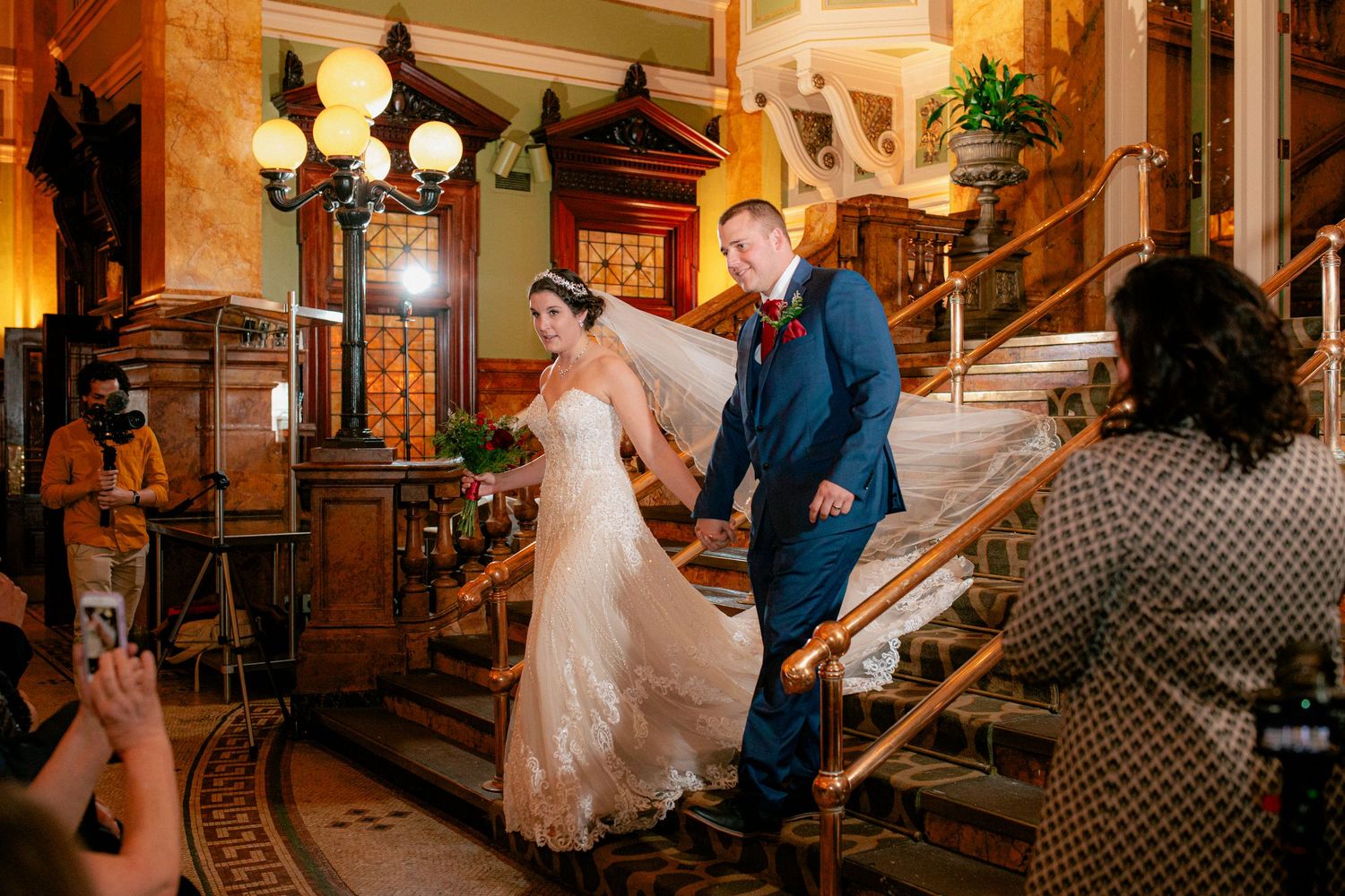 Bride in lace gown and groom in blue suit make grand entrance down ornate staircase at wedding.