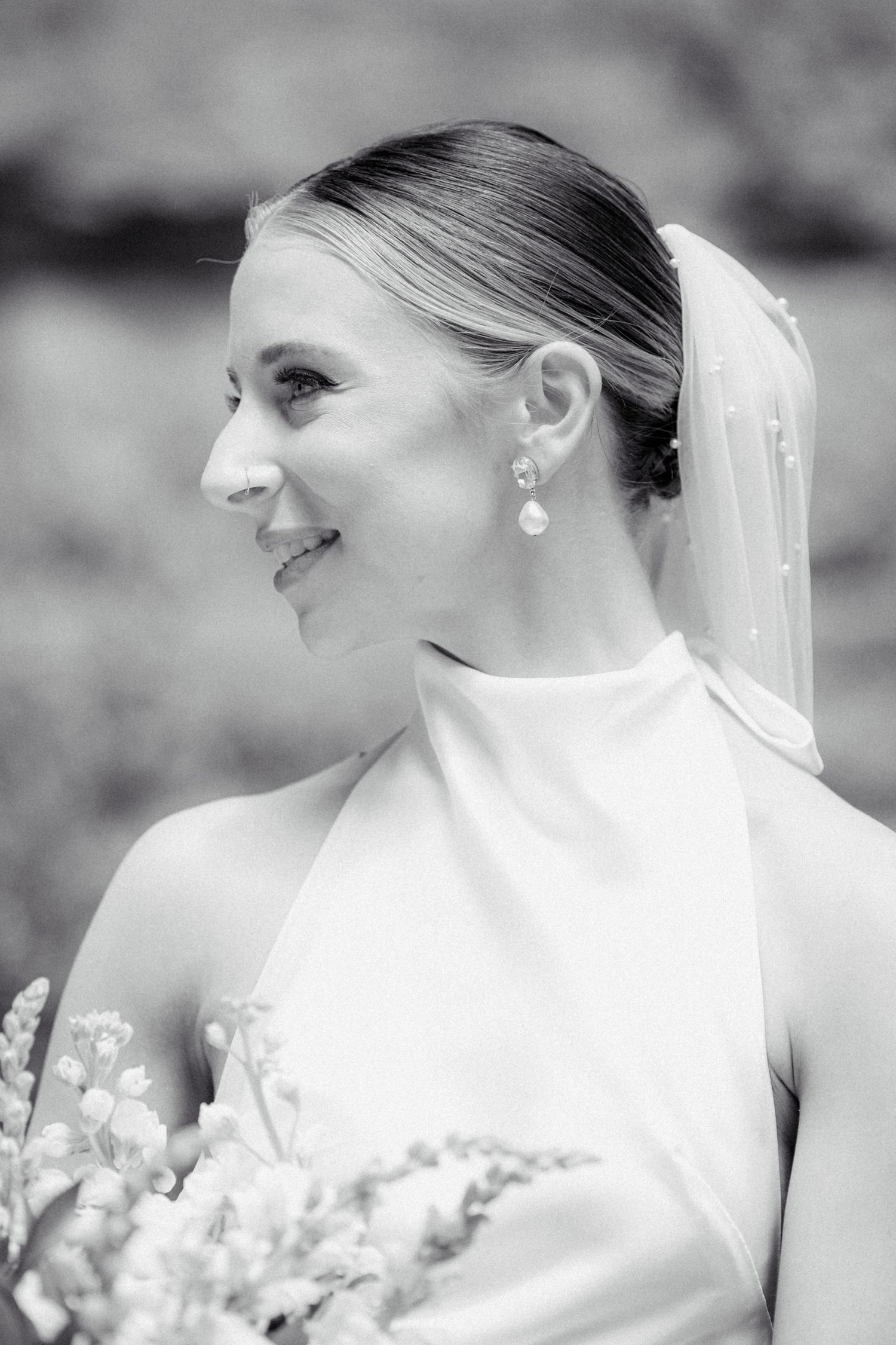 Black and white portrait of smiling bride wearing pearl earrings, veil, and halter wedding gown holding bouquet.