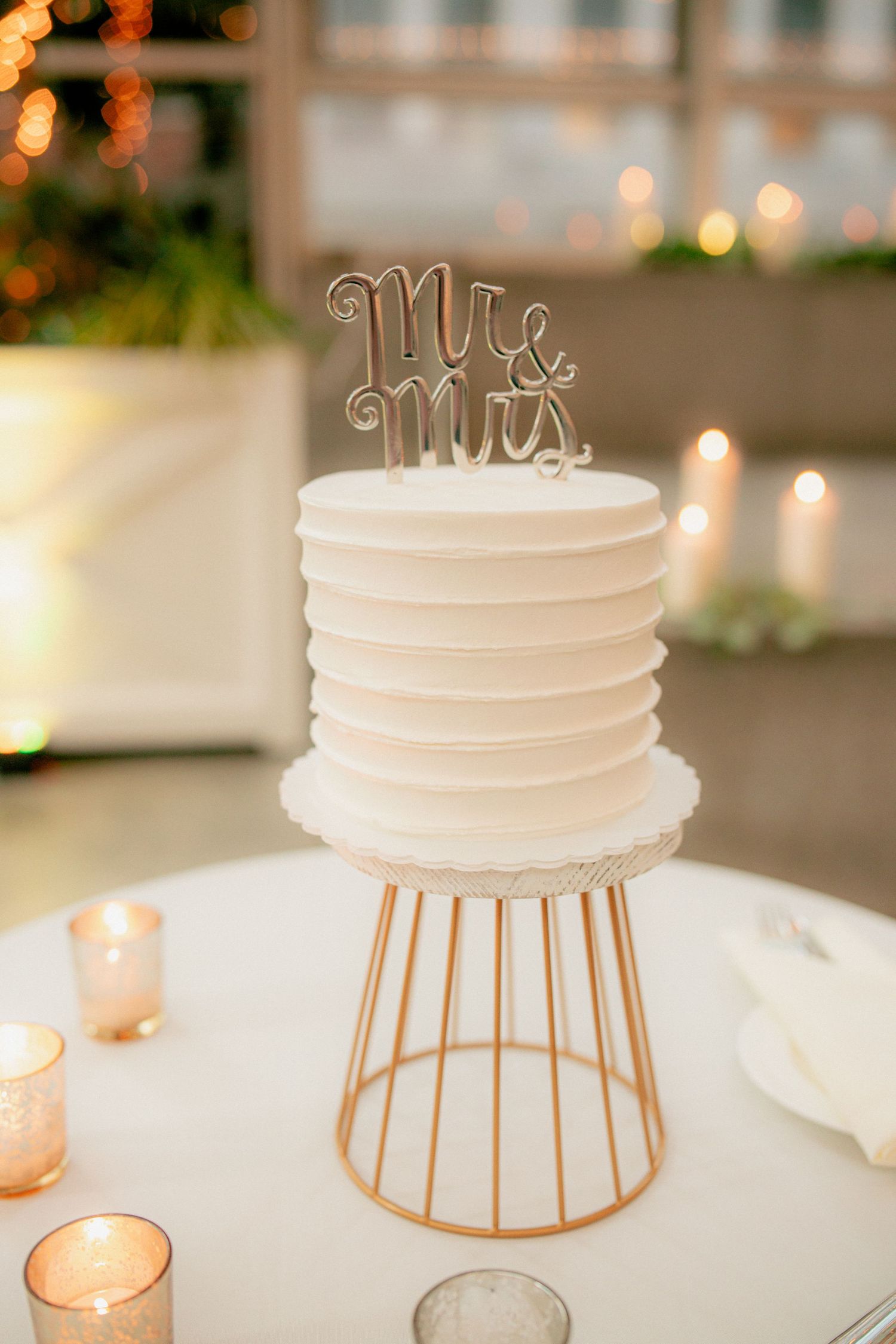 Single-tier white ribbed wedding cake with Mr. and Mrs. topper displayed on a gold geometric stand with votive candles.