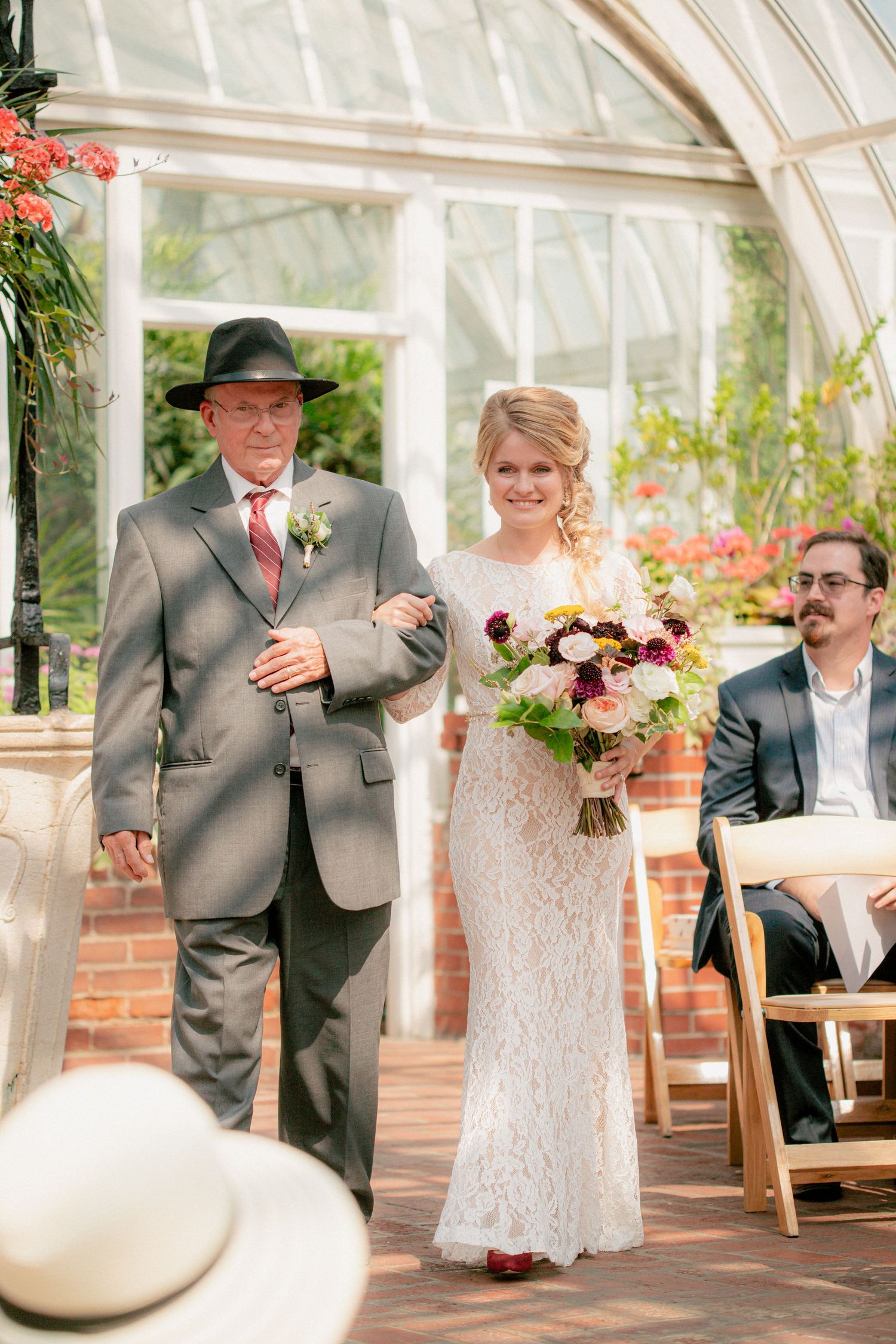Bride walks down the aisle with her father in a sunlit greenhouse wedding ceremony.