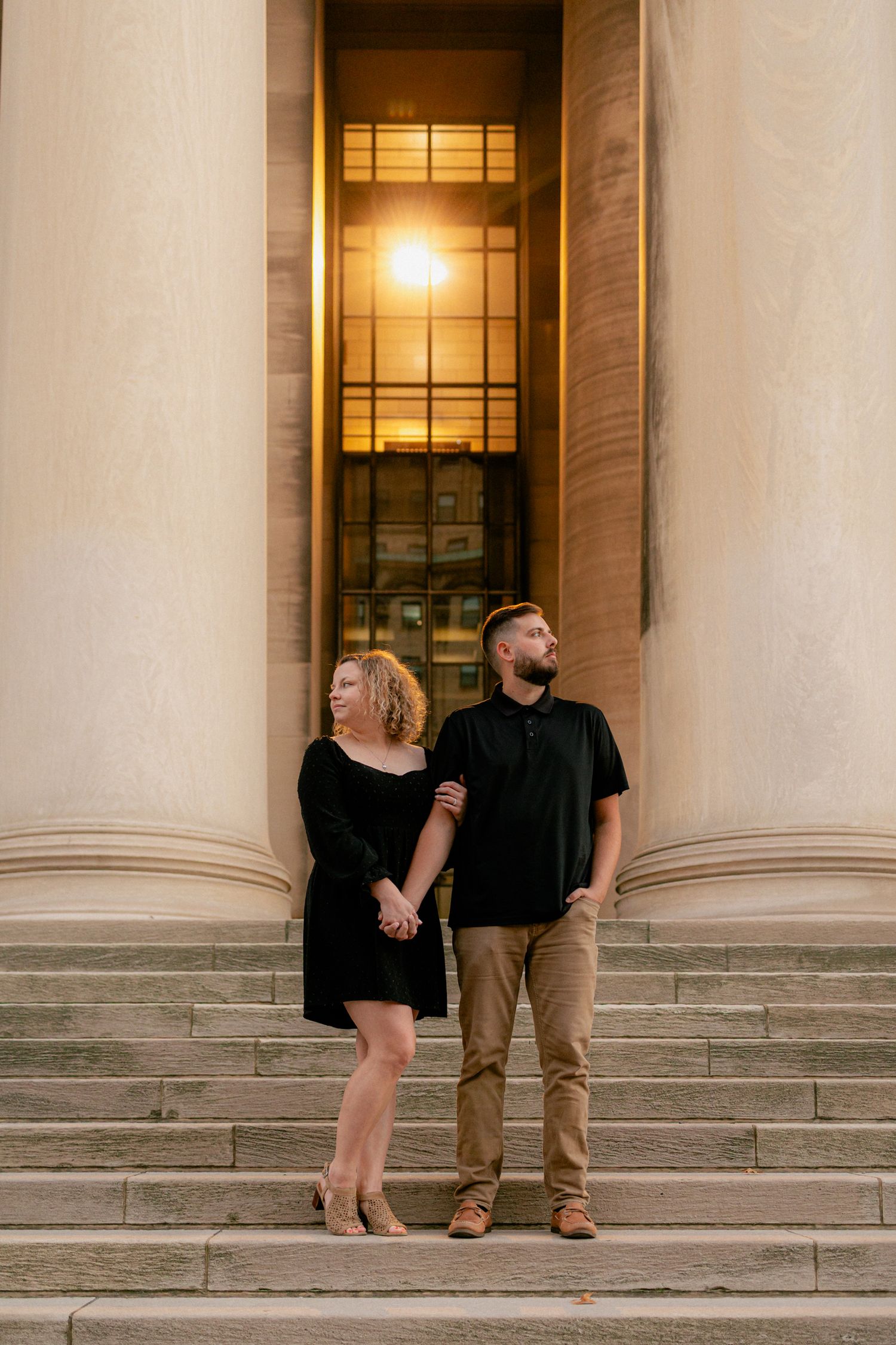 Couple holding hands on steps of grand building with tall columns, dressed in matching black outfits.