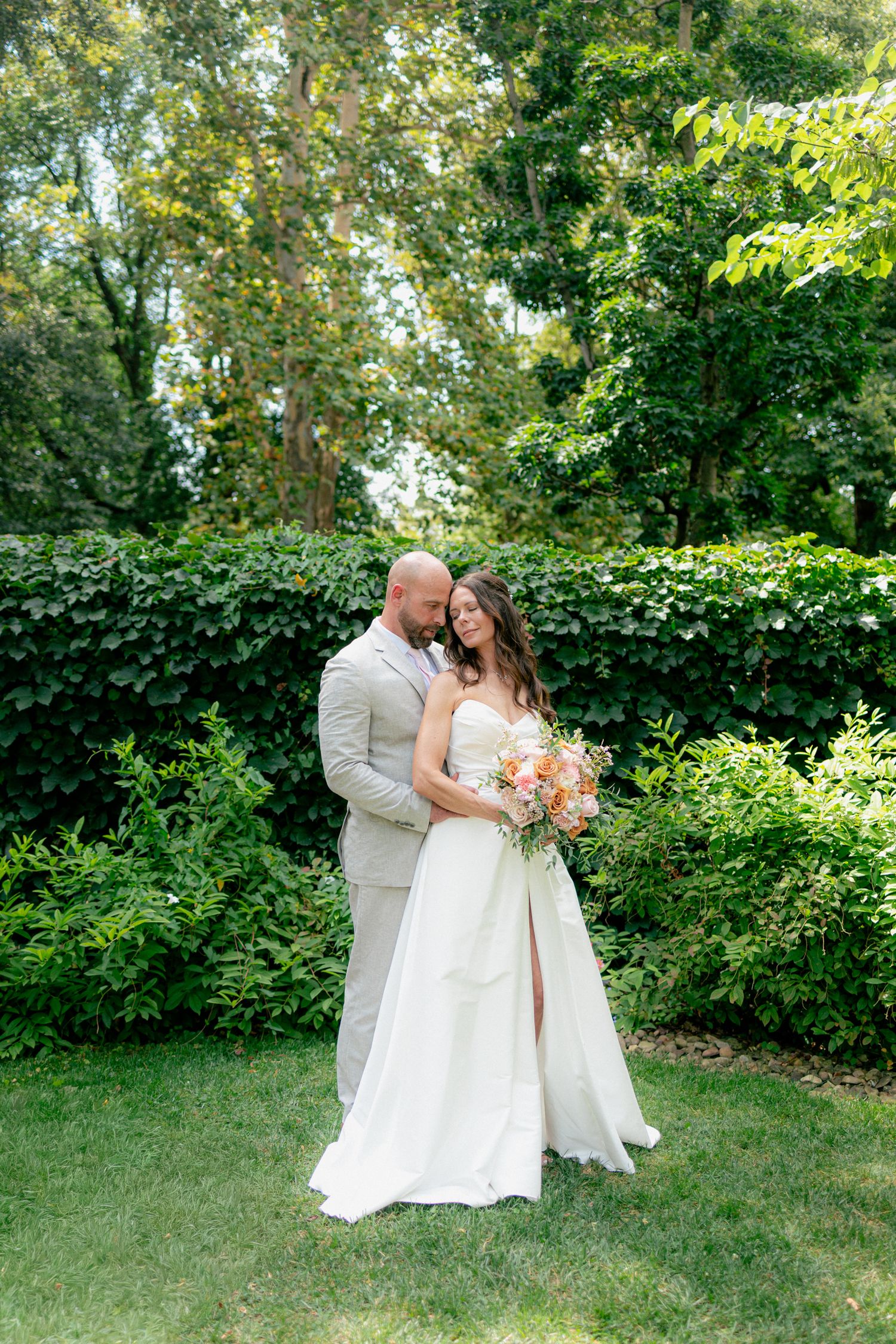 Bride and groom embracing in a lush green garden, bride in white strapless gown holding a peach bouquet.
