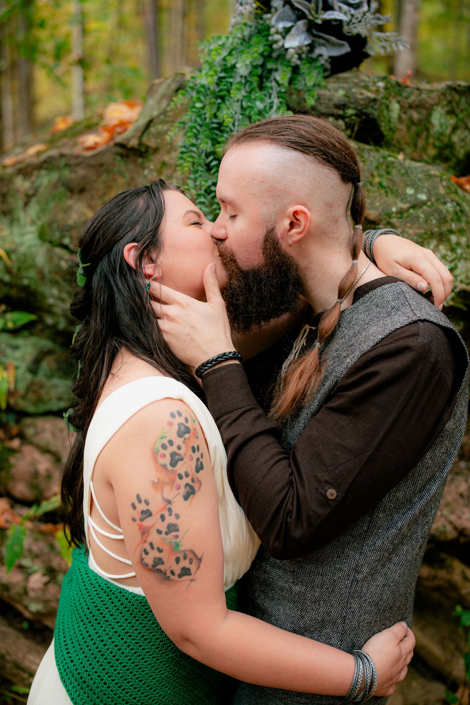 Tattooed bride with green dress kisses bearded groom near mossy rocks in autumn forest wedding.