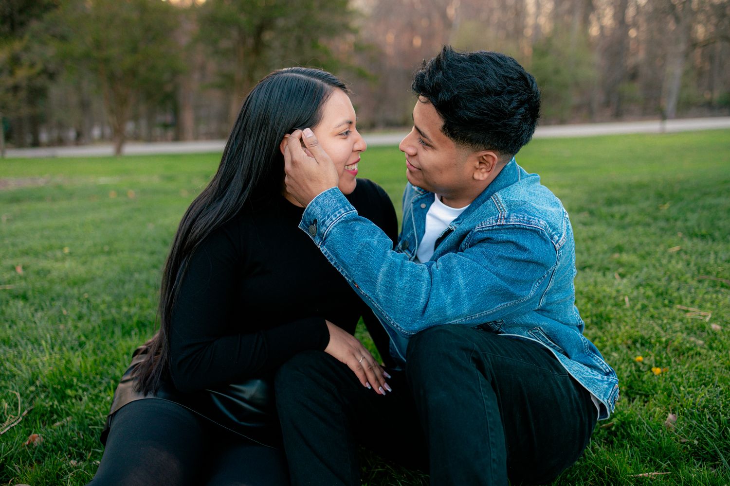 Couple sitting on green grass in a park, man tenderly cupping womans face while gazing at her.