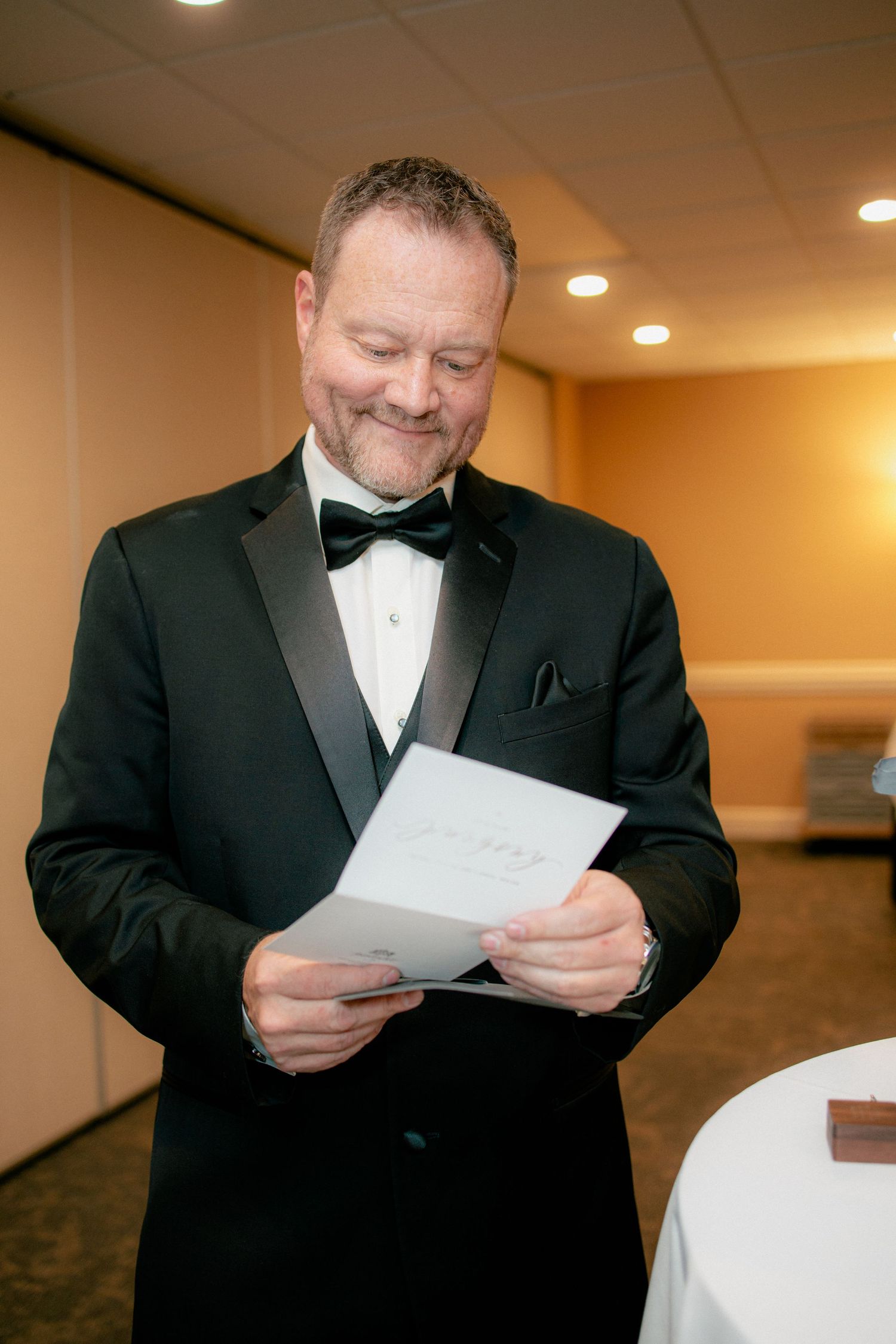 Groom in black tuxedo smiling while reading a wedding card in a banquet hall.