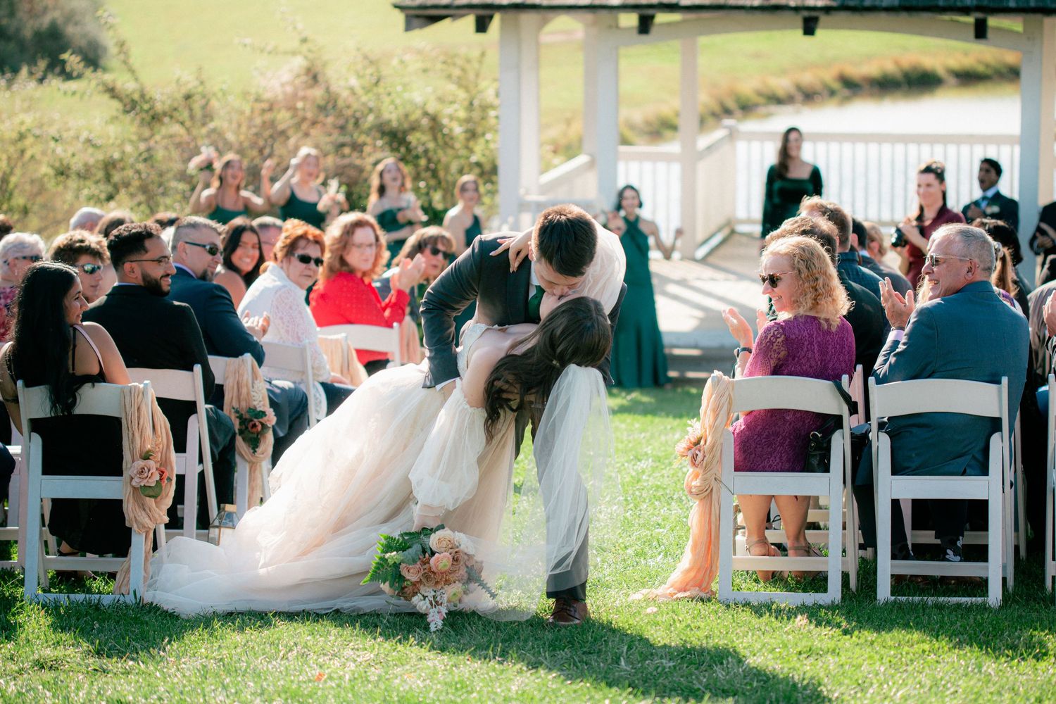 Groom dips bride for a romantic kiss on the grass aisle surrounded by seated wedding guests.