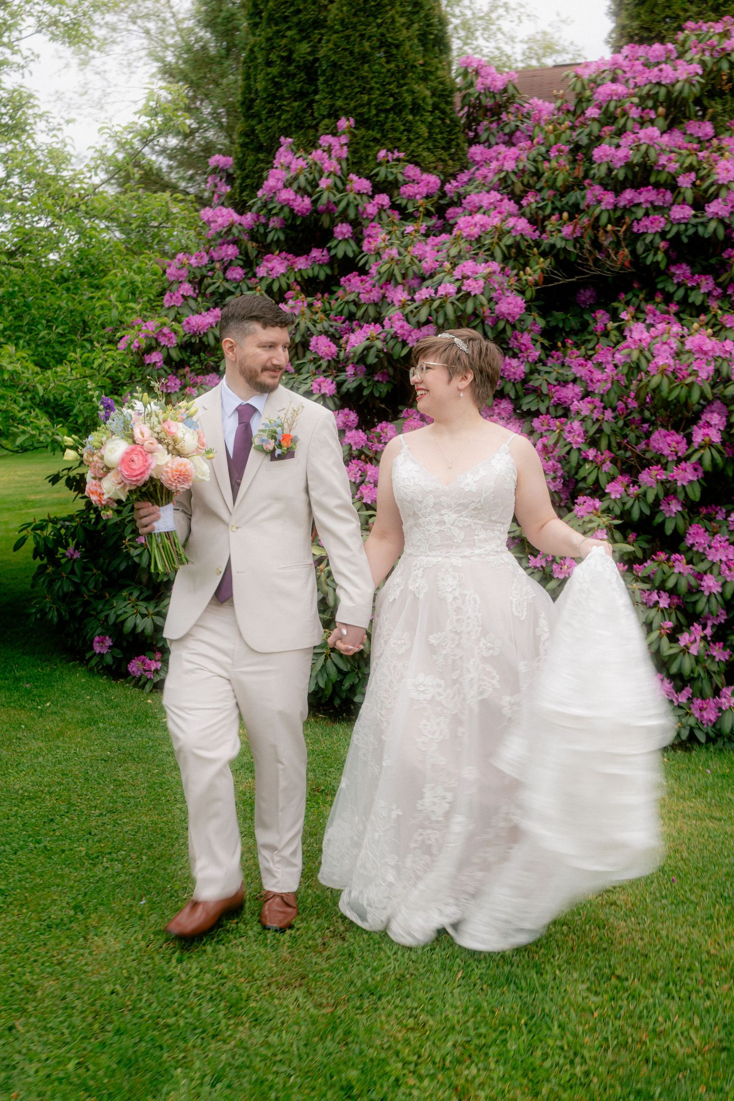 Bride and groom walk hand-in-hand on green lawn before blooming pink rhododendron bushes in wedding attire.