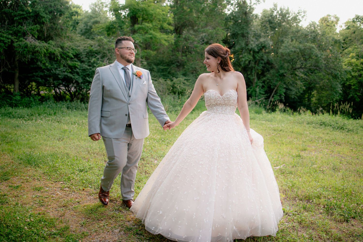 Bride in ballgown and groom in gray suit walk hand-in-hand through a lush green outdoor wedding setting.