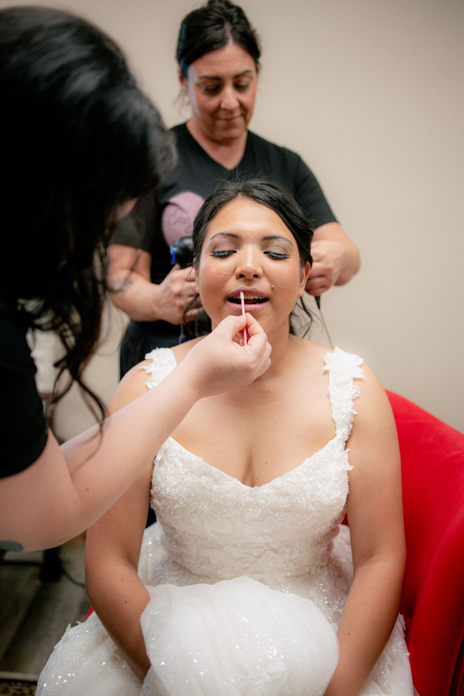 Bride in white lace wedding dress getting makeup and hair done by two stylists before her wedding.