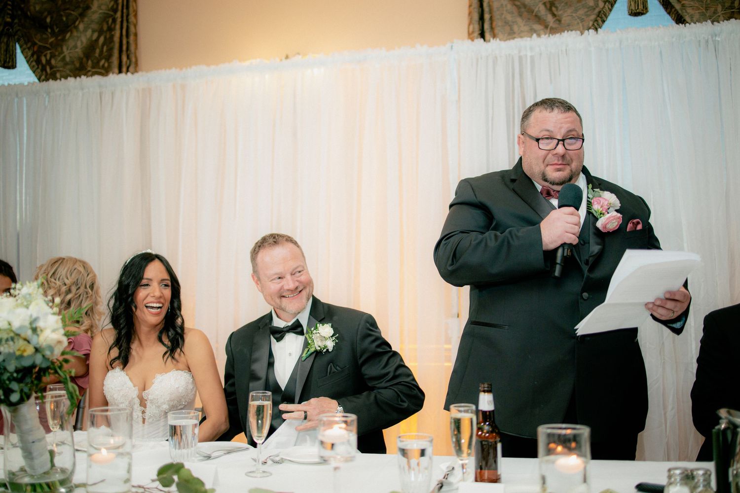 Best man delivering wedding speech to laughing bride and groom at elegant reception.