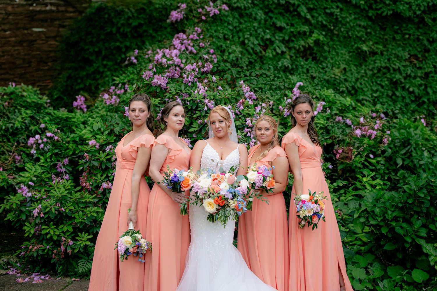 Bride in white gown poses with four bridesmaids in coral dresses holding colorful bouquets before a flowering green hedge.