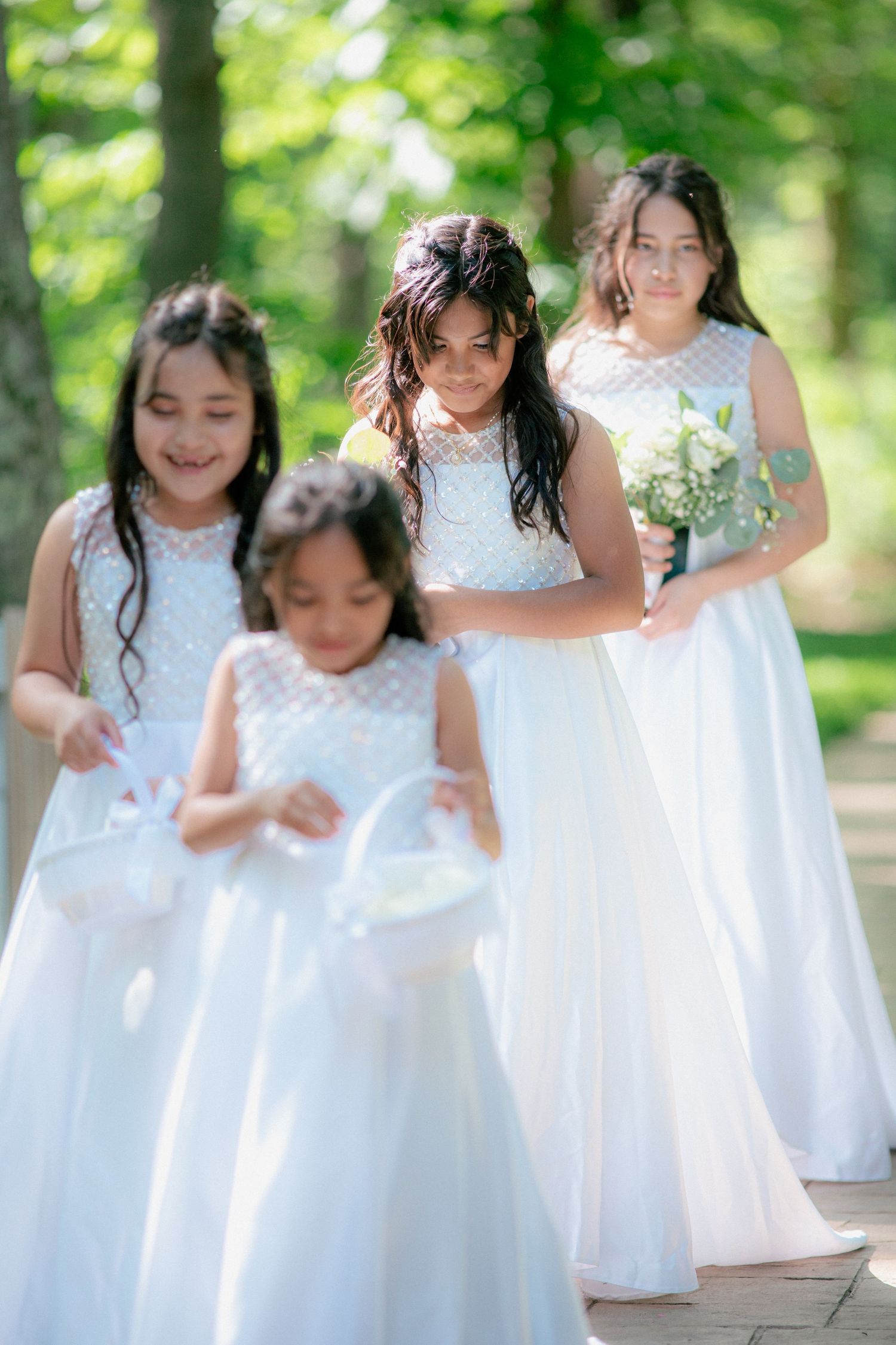 Four young girls in white dresses walk outdoors, with flower girls carrying baskets at a wedding ceremony.