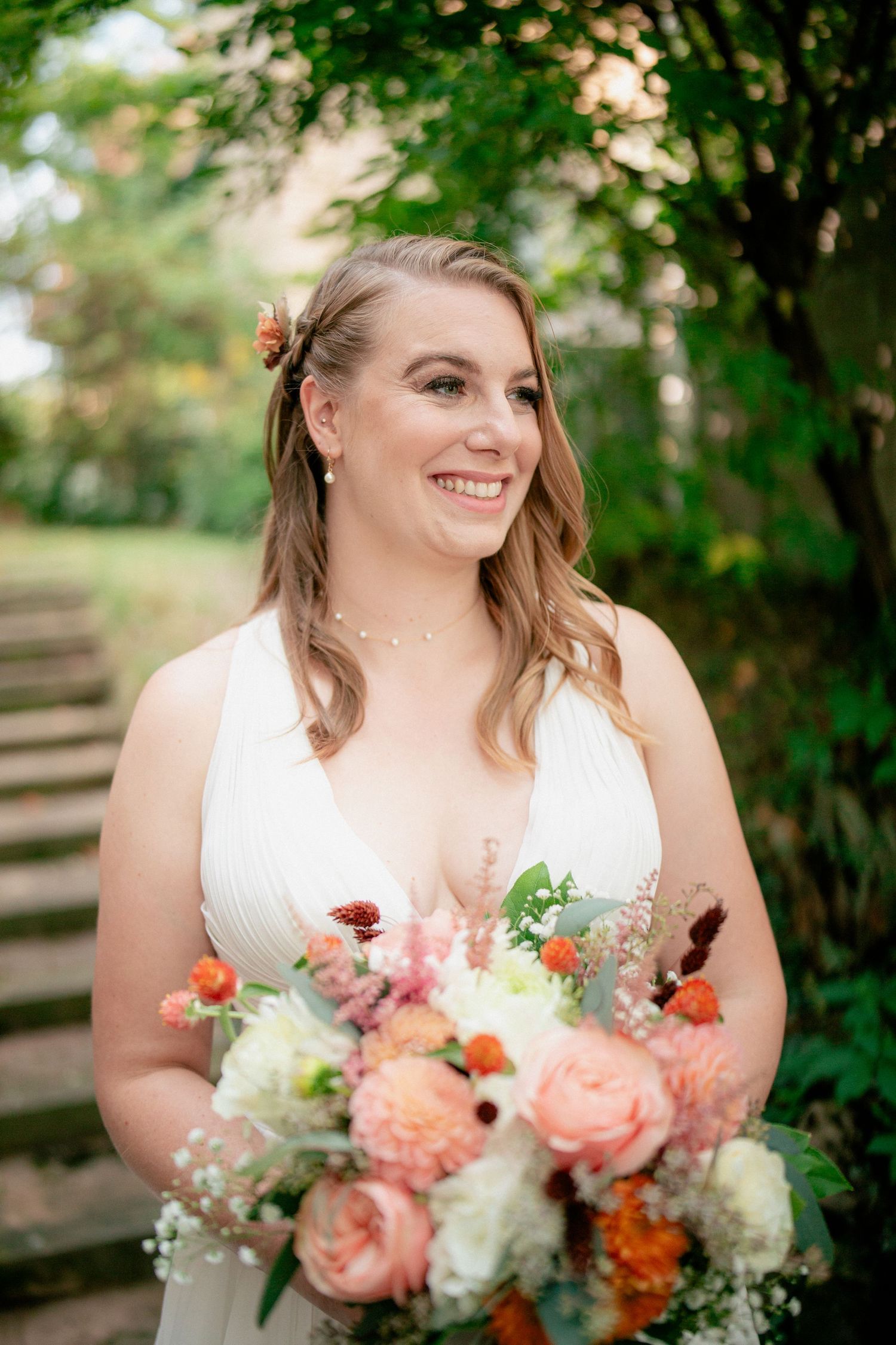Smiling bride in white V-neck gown holds colorful bouquet with flowers in her hair outdoors.