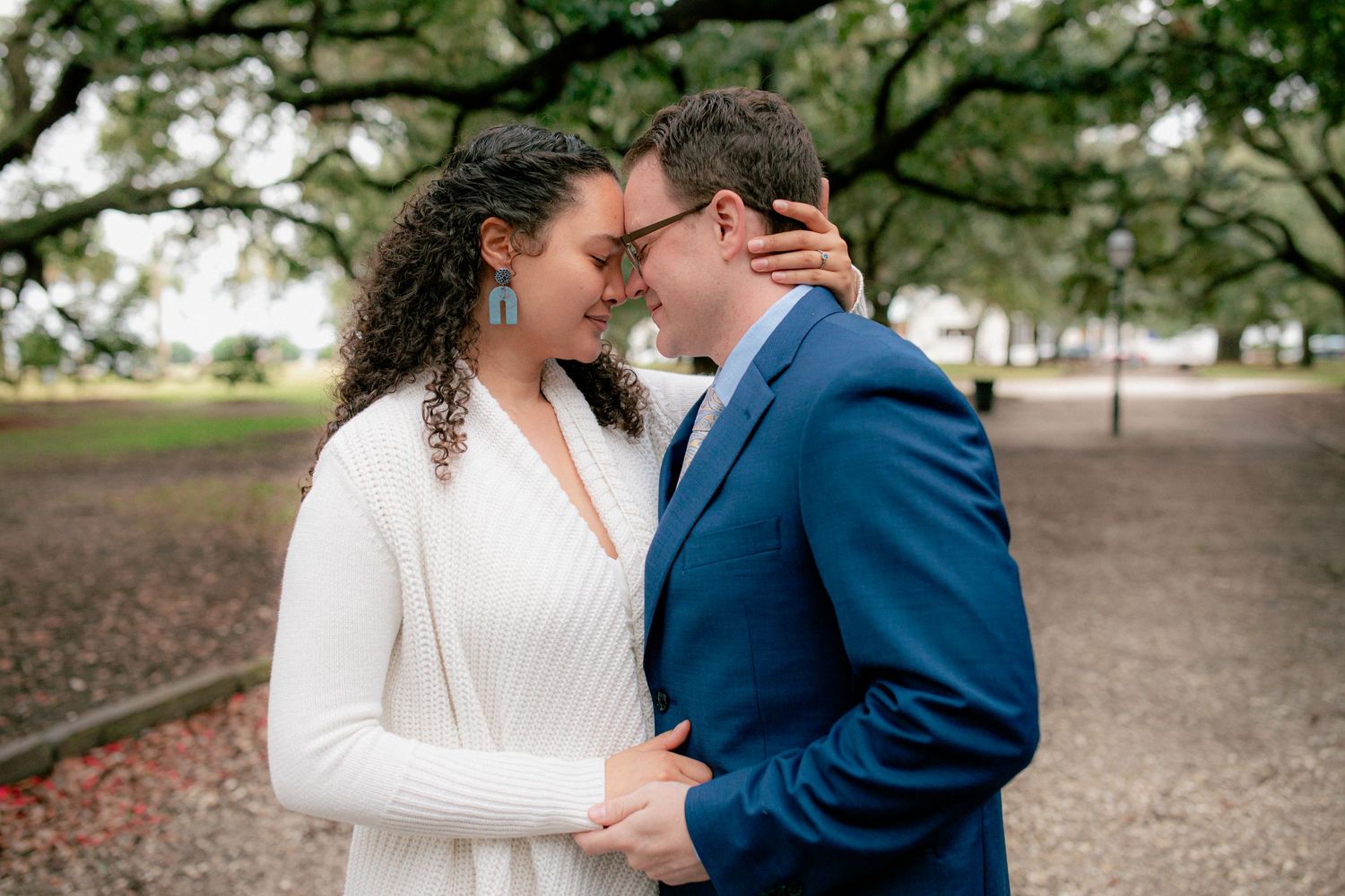 Bride and groom embraces tenderly under a canopy of oak trees, woman in white sweater and man in blue suit.