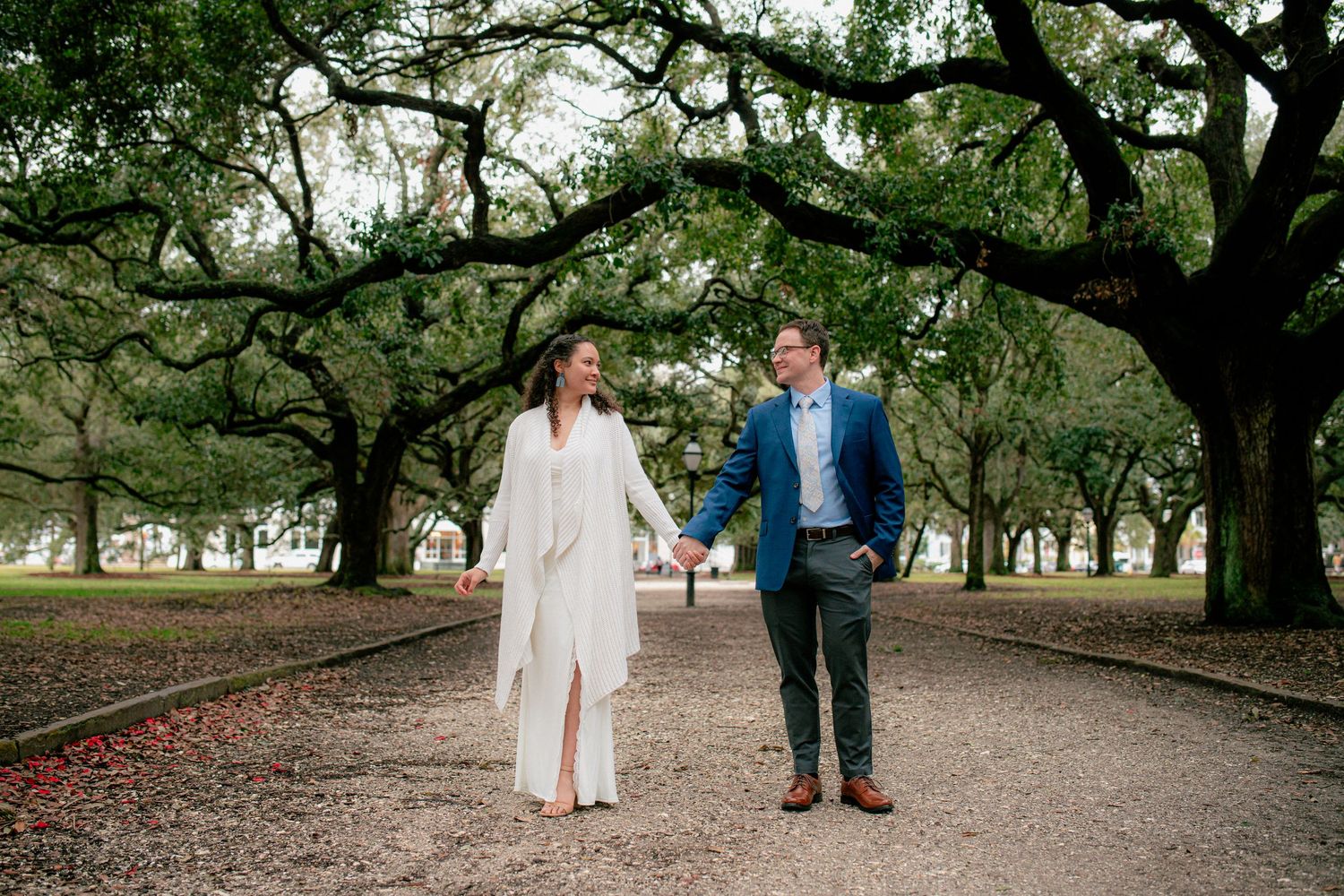 Happy couple walks hand-in-hand down a scenic oak-lined path, woman in white dress and man in blue suit.