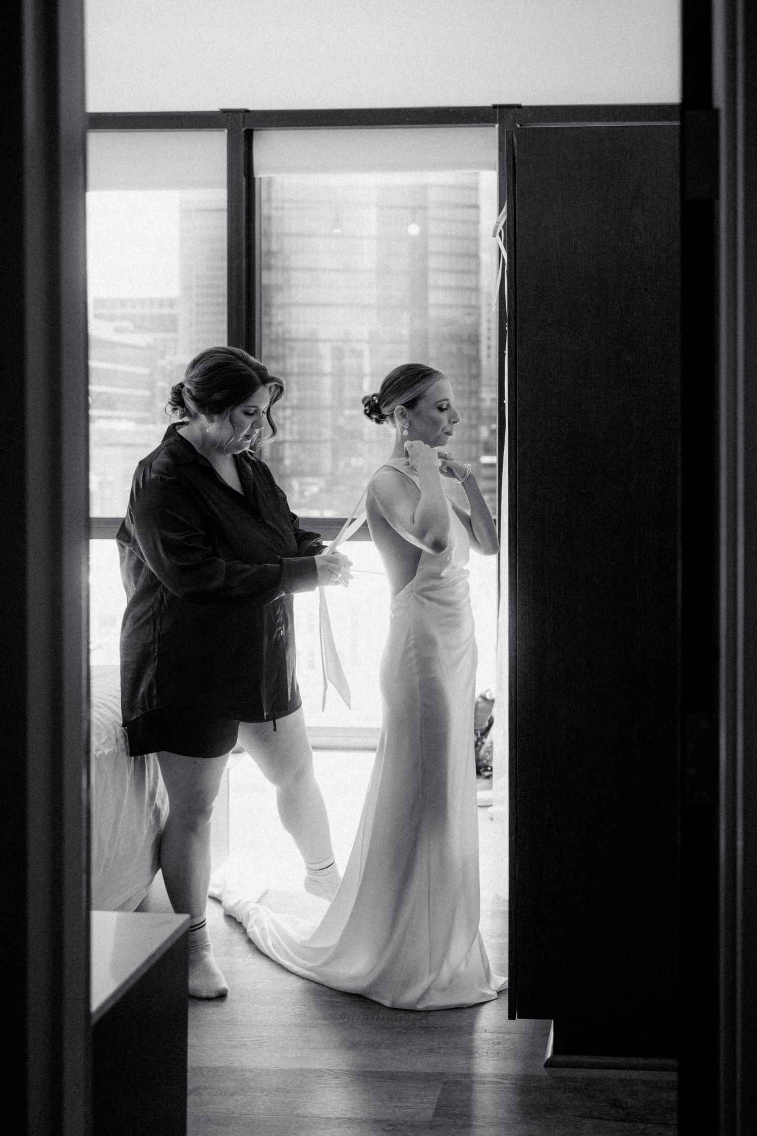 Black and white photo of bridesmaid helping bride with dress before wedding ceremony.