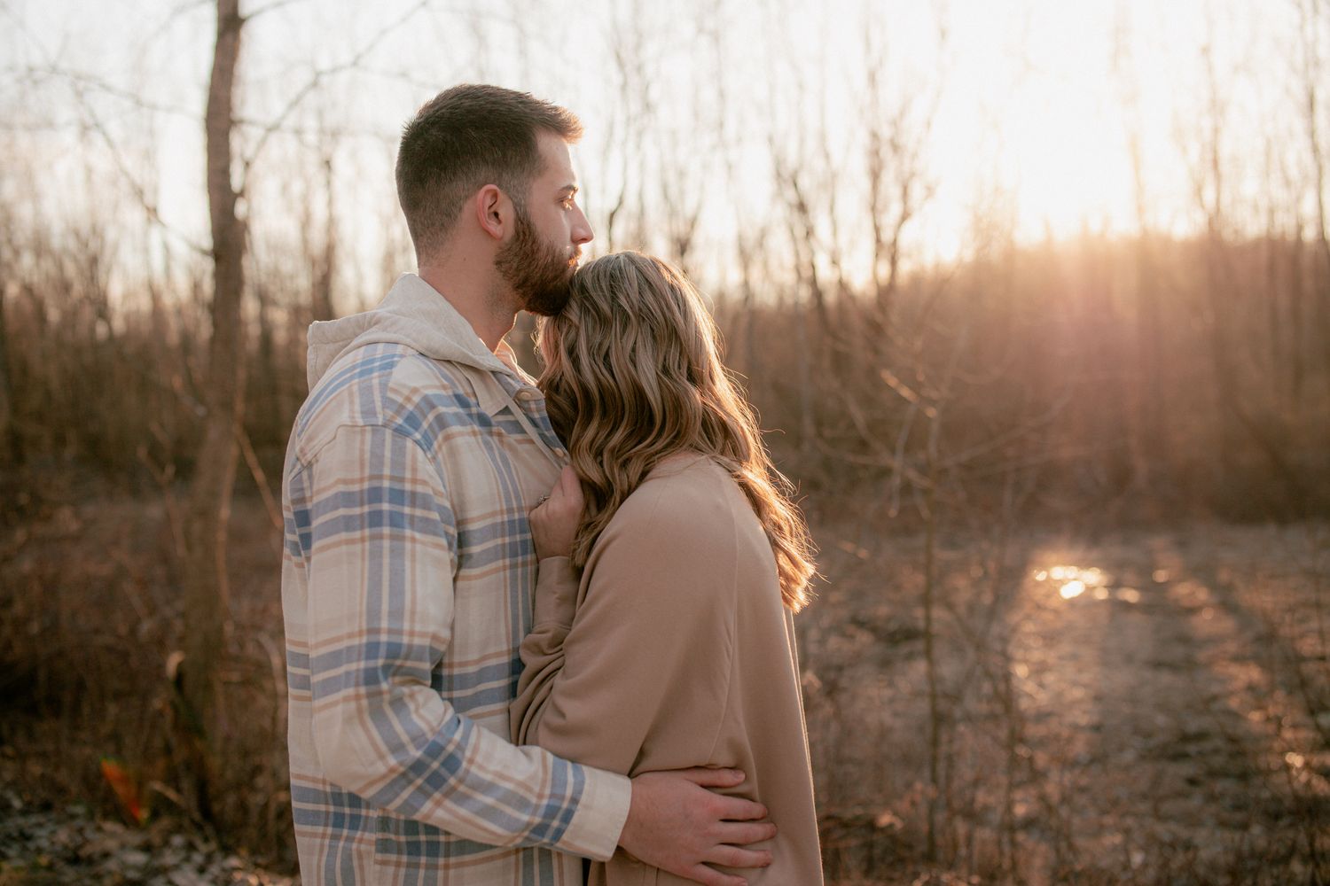 Couple sharing a tender sunset kiss outdoors surrounded by bare winter trees in warm golden light.