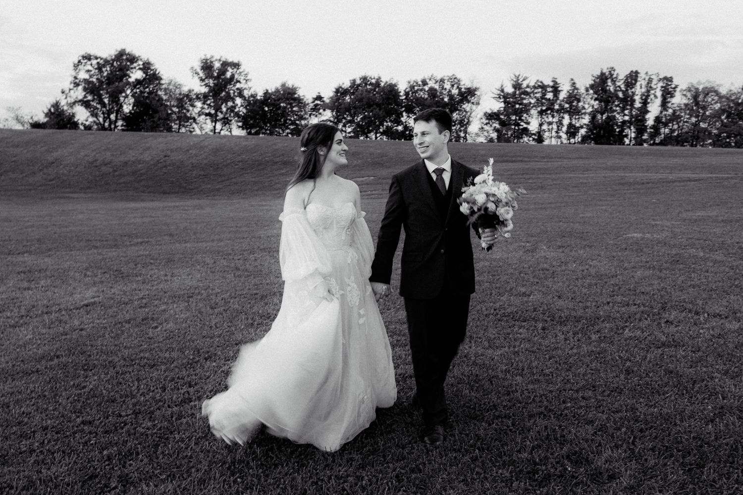 Black and white photo of smiling bride and groom holding hands walking across a lush field.