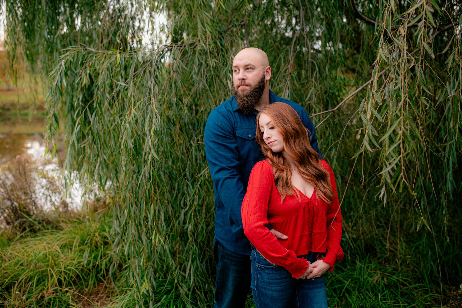 Bearded man and redhead woman standing under weeping willow by pond.