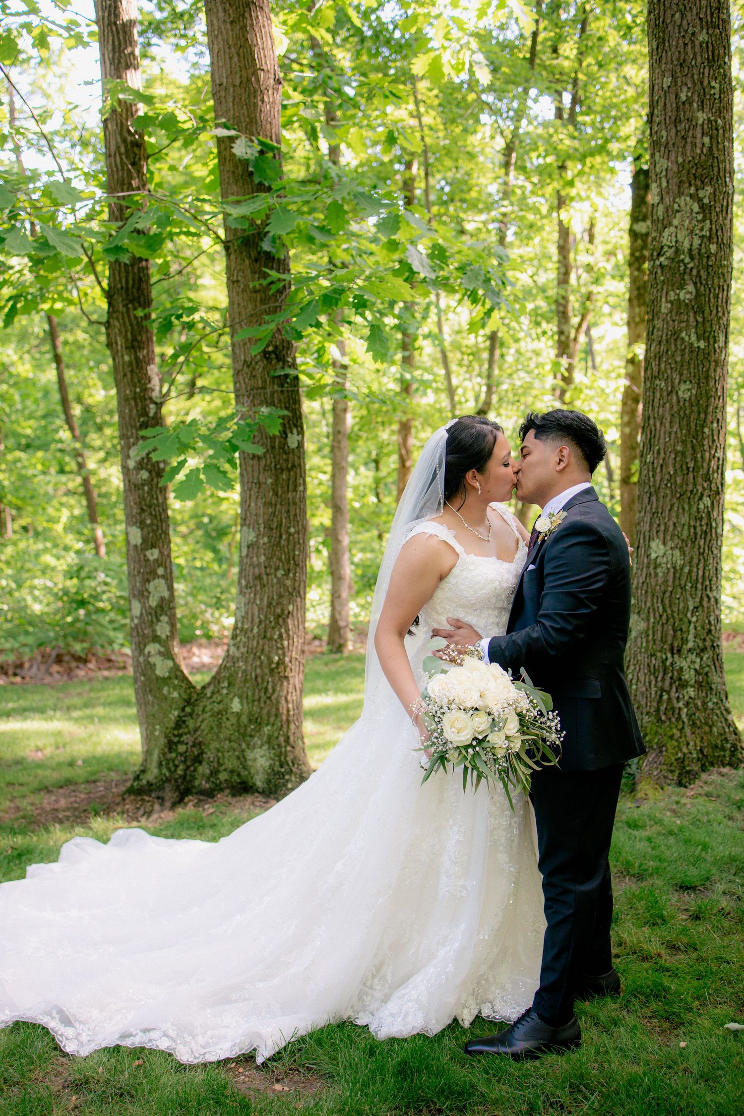 Bride and groom share a tender kiss surrounded by lush green trees, her long lace gown trailing gracefully on the ground.