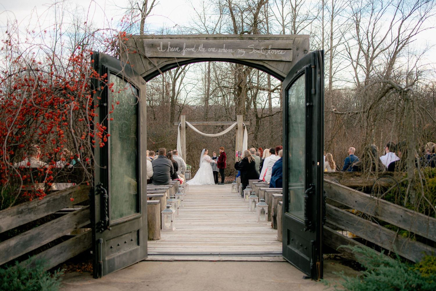 Wedding ceremony viewed through rustic wooden doors, guests seated along an outdoor aisle.