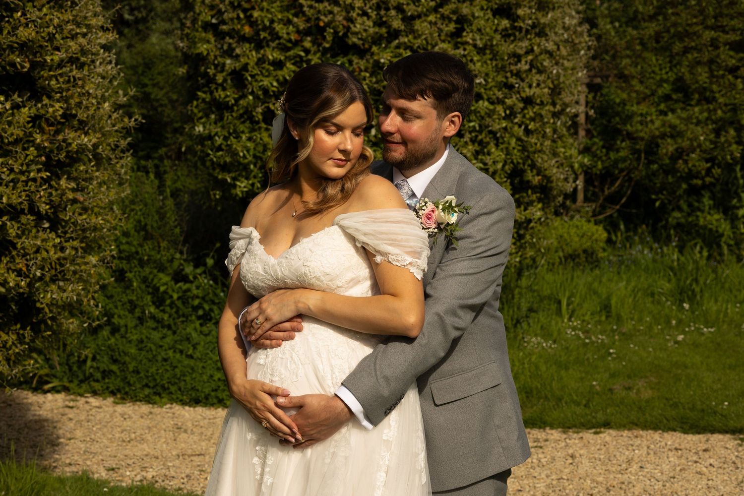 An image of a groom stood behind the bride holding her bump as they turn their heads towards one another