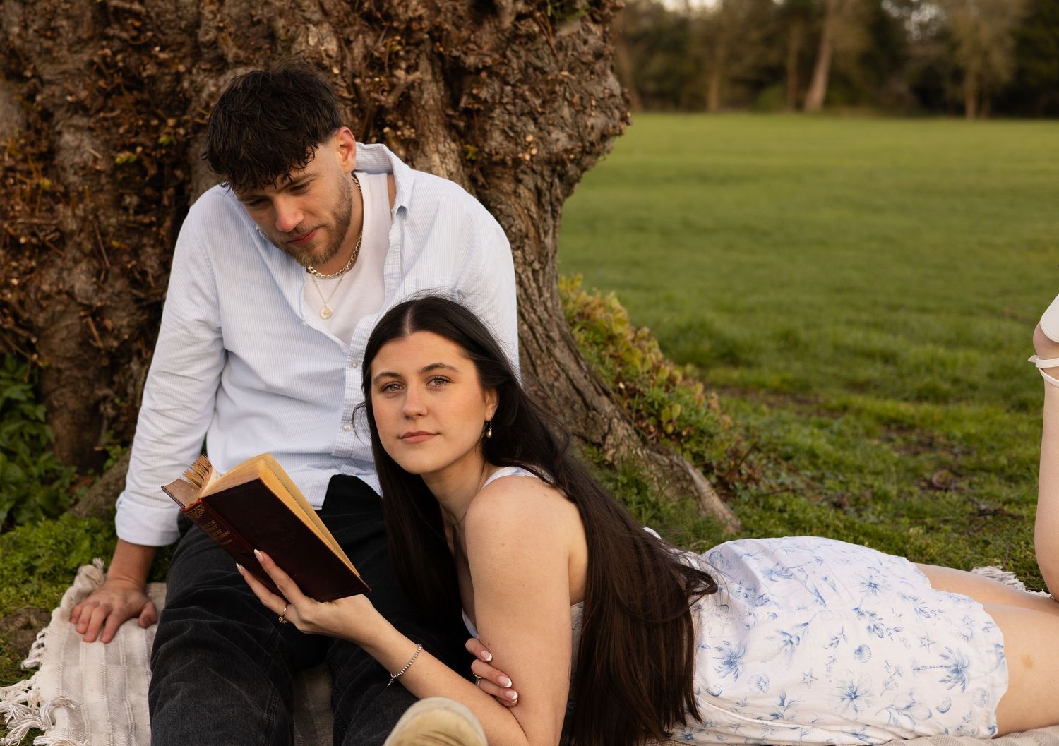 A couple sat on a picnic blanket. The man is sat against a tree and the woman is led on her, holding a book while looking into the camera.
