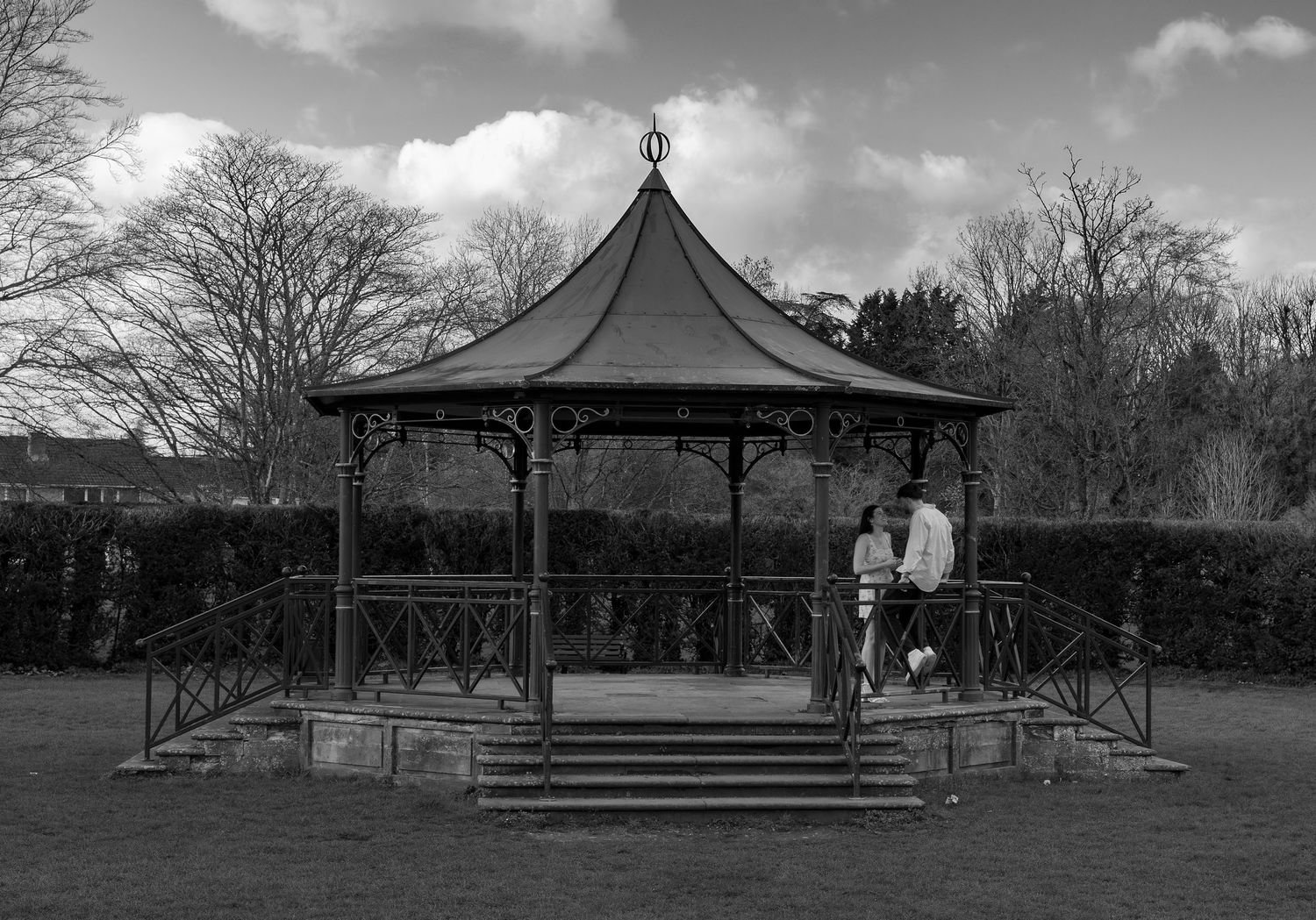 A black and white image of a couple in a bandstand. The man is sat on the railing and the woman looks up at him.