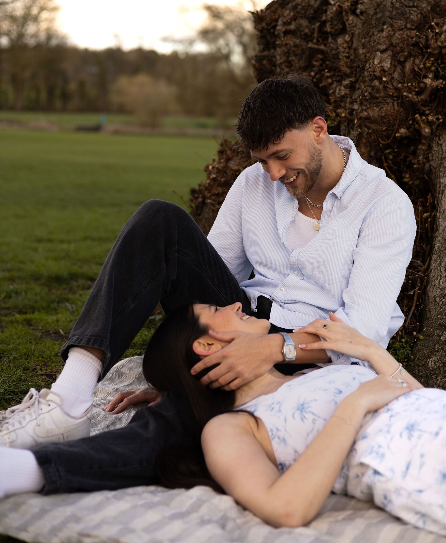 A man sat on a picnic blanket leaning against a tree. A woman is led with her head in his lap looking up at him. He holds her face,