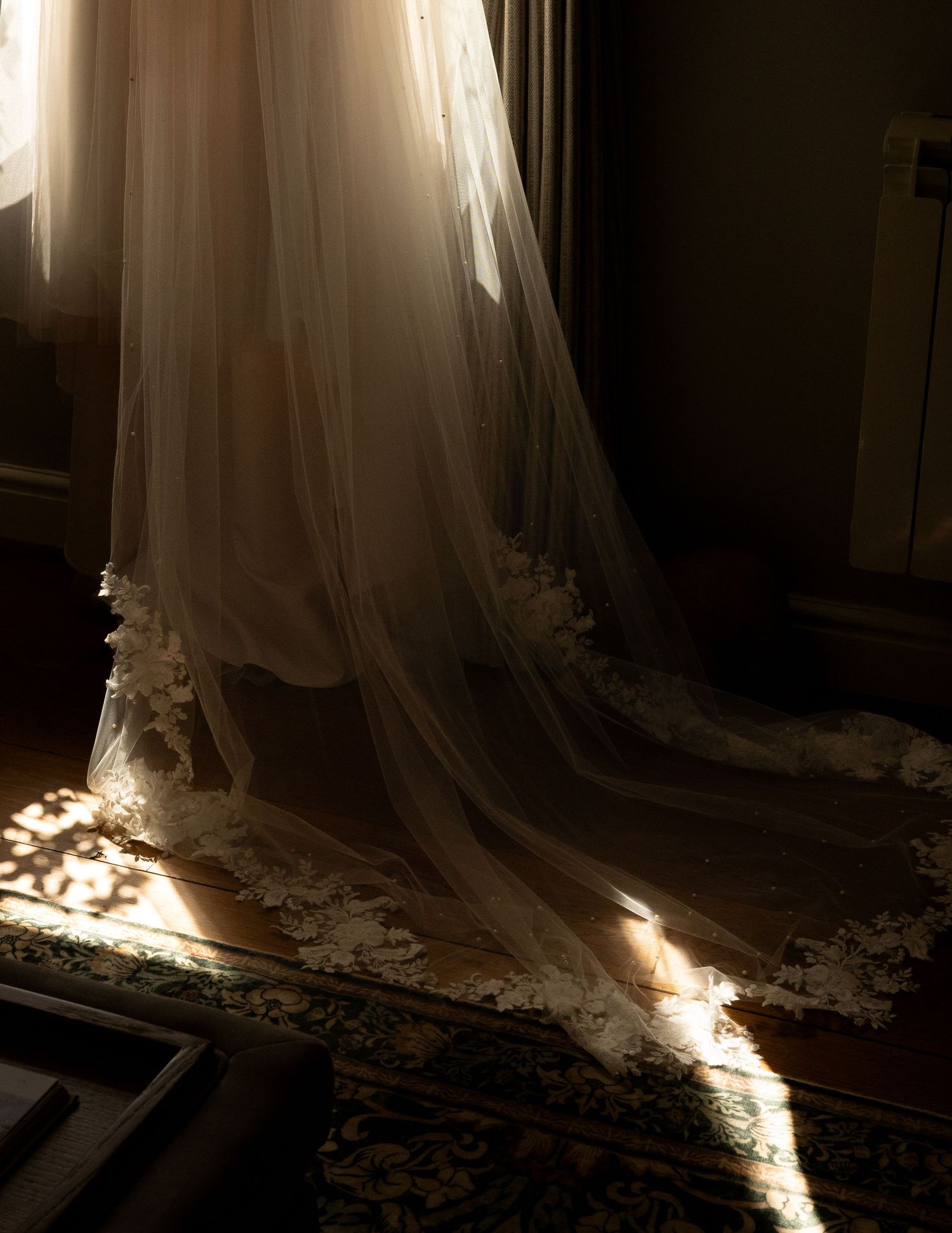 The bottom of a wedding dress with a veil hanging in the window of a dark room with the light shining through