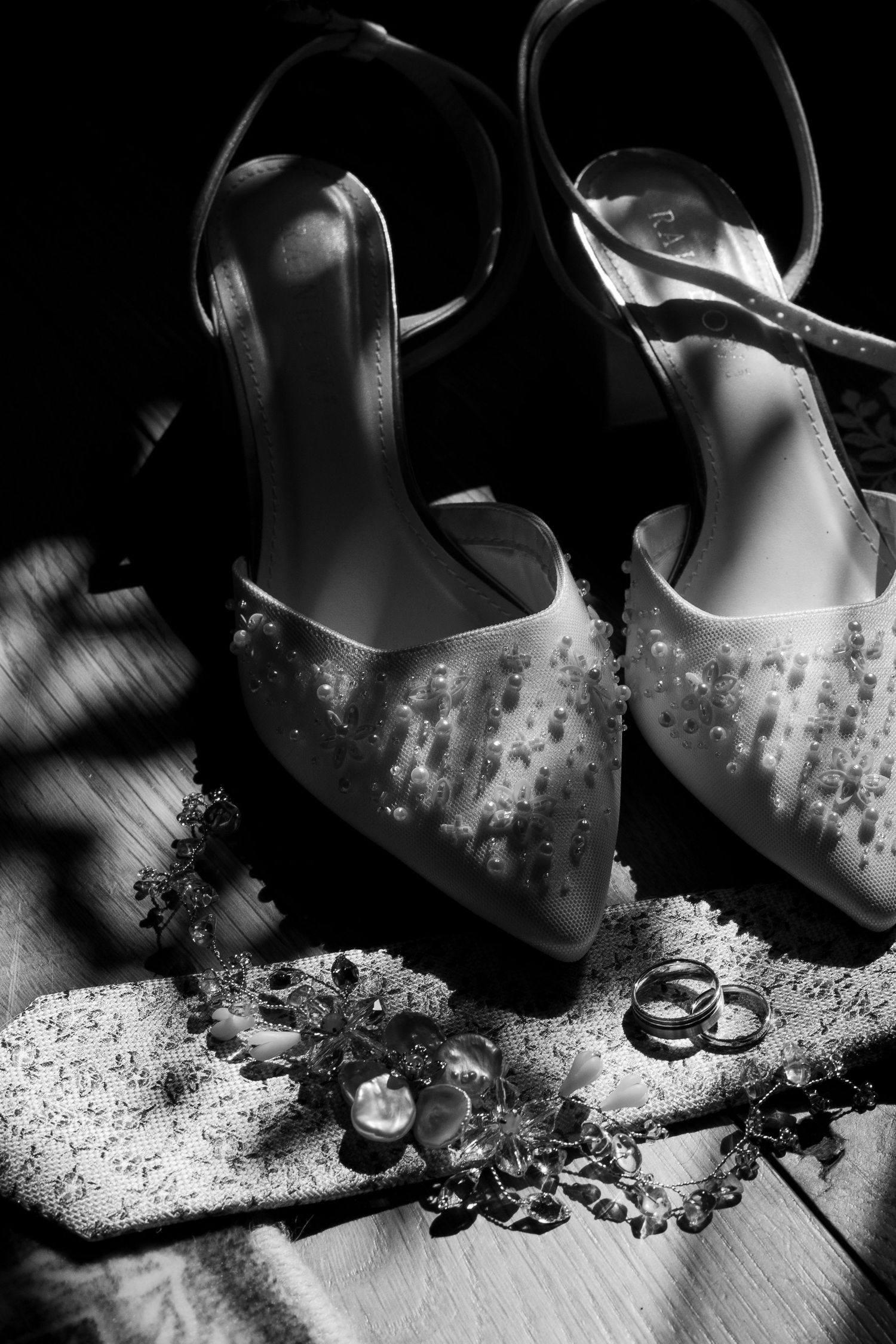 A black and white image of some wedding items placed on the floor in sunlight. The items include, rings, a tie, shoes and a hair piece
