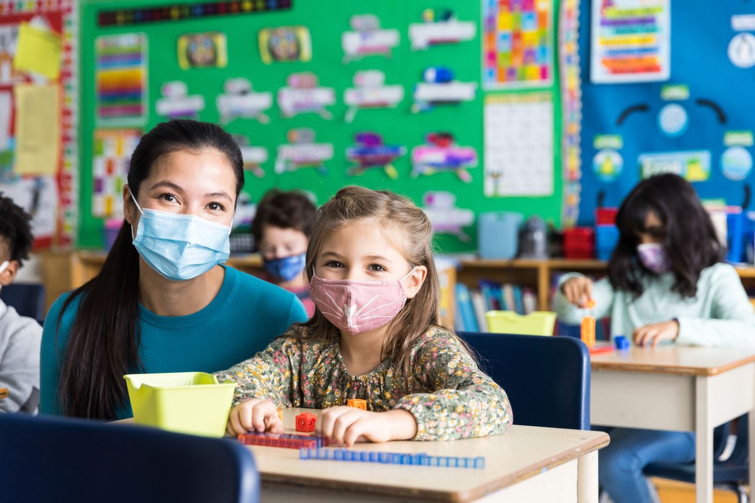 A teacher and a student sit smiling in a colourful classroom, both are wearing masks.