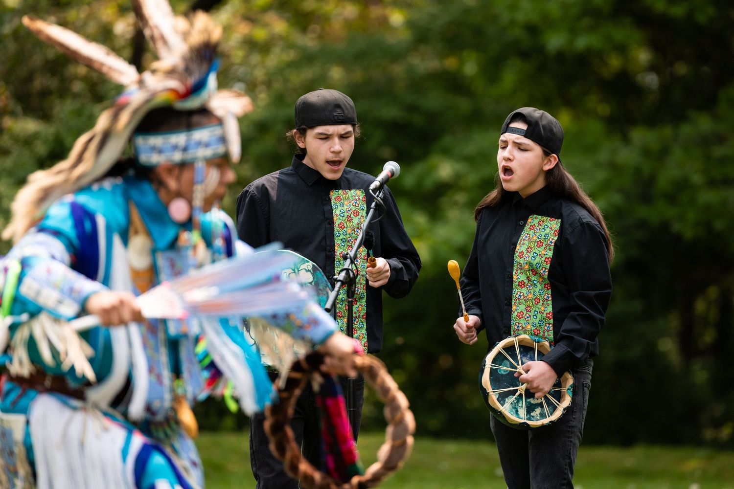 Two young indigenous men sing and play drums while a sun dancer in regalia dances