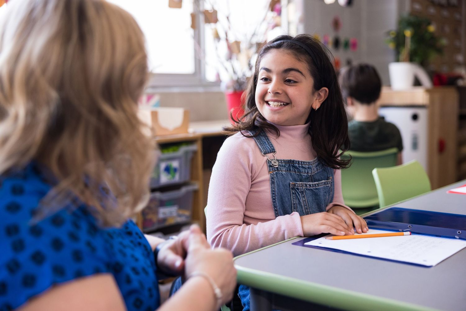 A young female student in overalls smiles at a teacher while they sit at her desk. Commercial lifestyle photography, kids, families.