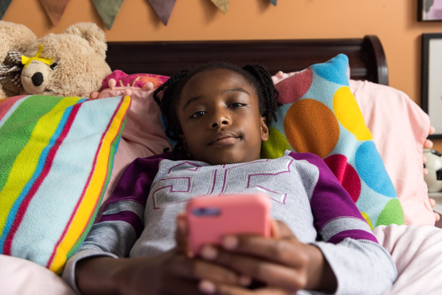 A young black girl lays on bed on her phone, looking sad. Photography for tech, education, law.