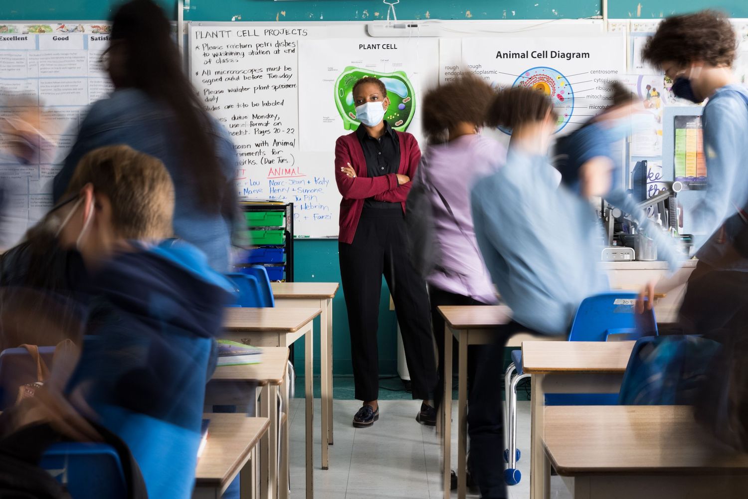 A teacher with a mask on stands in front of a crowded classroom full of students in motion. Commercial lifestyle photography.