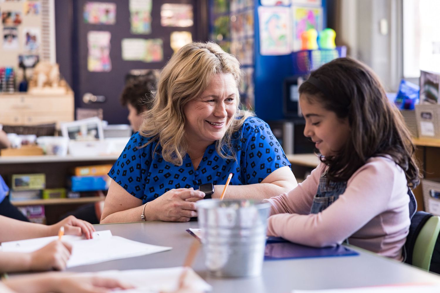A teacher smiles while helping a young student at her desk. Commercial lifestyle photography, back to school, school supplies, kids.