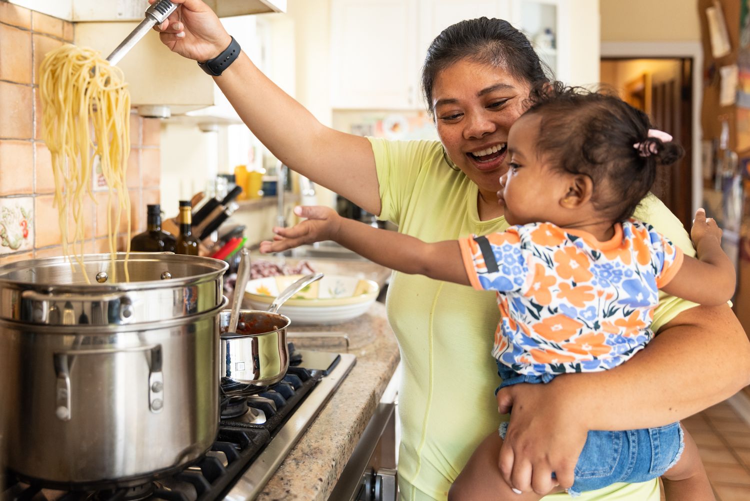 A young mother makes spaghetti in her kitchen while holding a young child. Commercial lifestyle photography.