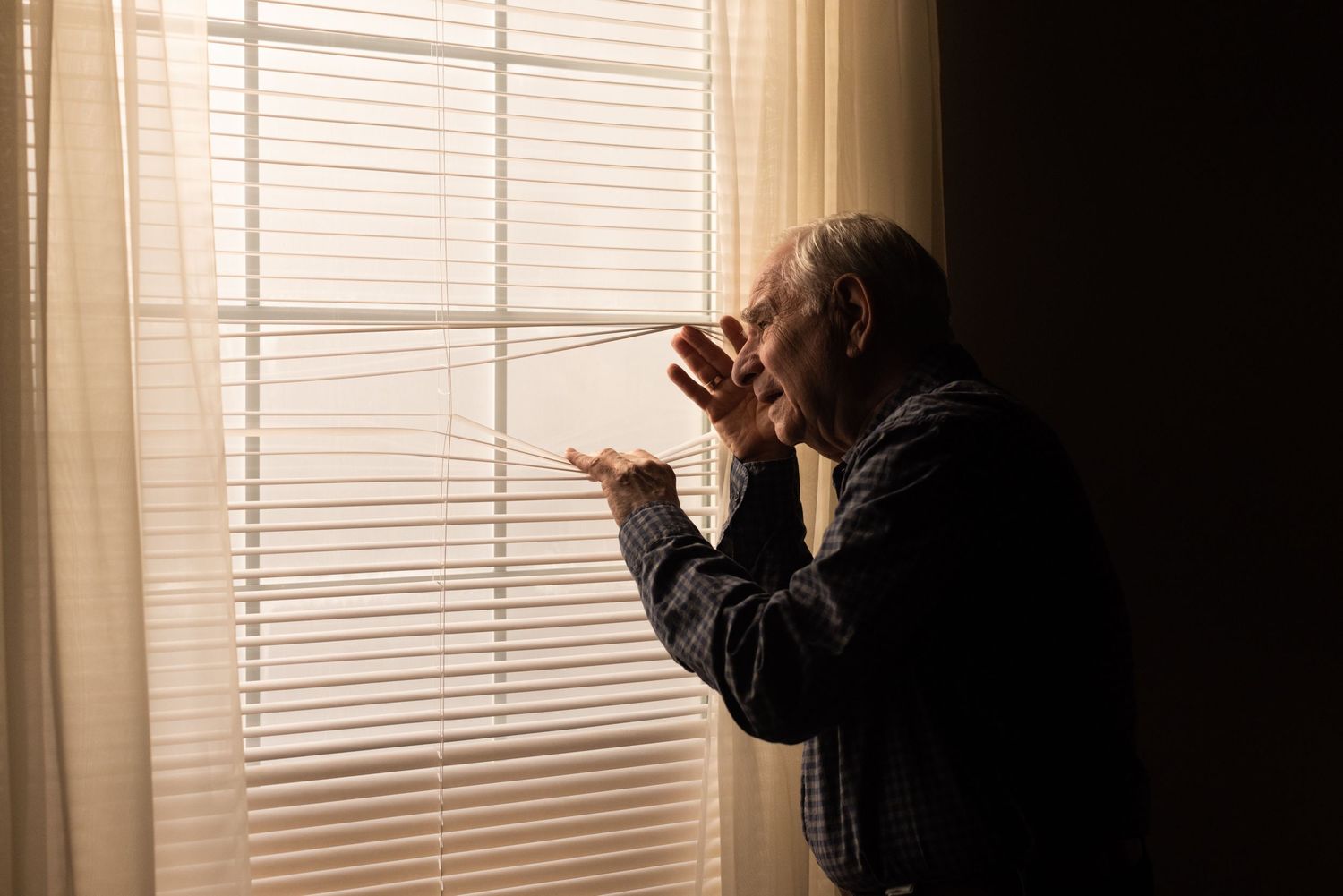 An elderly man peers out the blinds of a window, documentary commercial photography