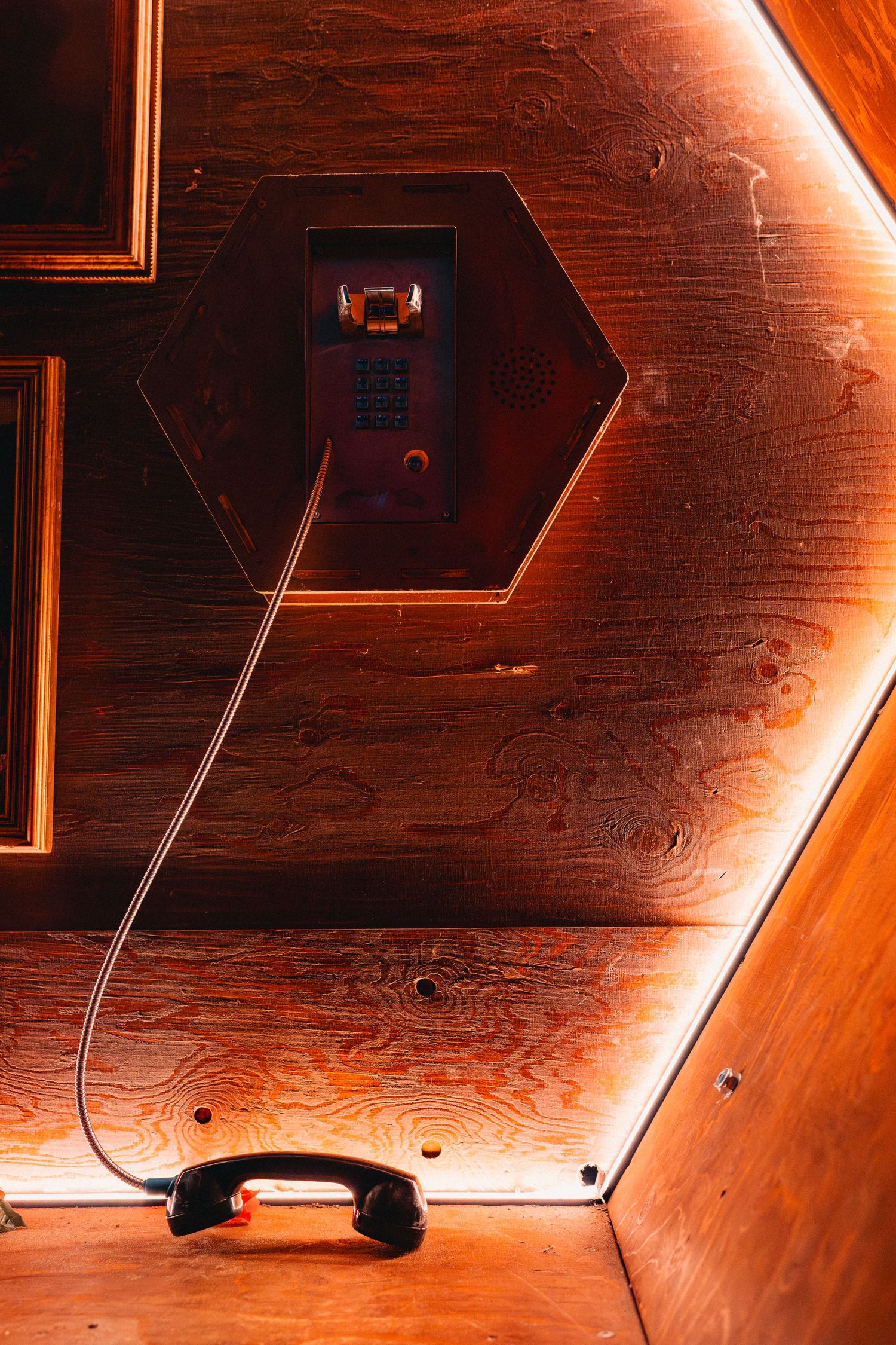 Vintage payphone mounted on warm wood-paneled wall with coiled cord hanging down in orange light.