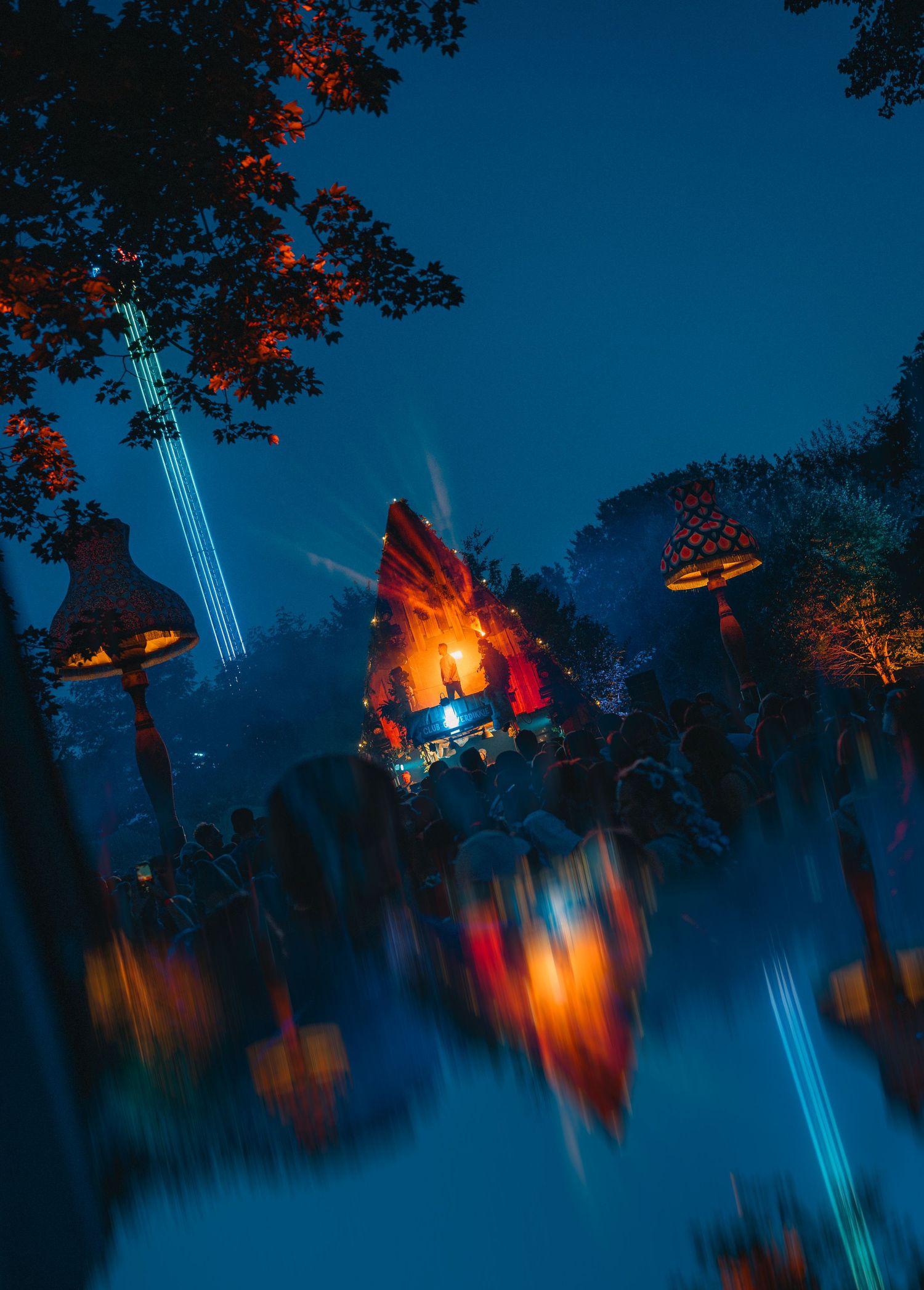 Blurry night festival scene with glowing fire installation, colorful lights, and crowds among trees at dusk.